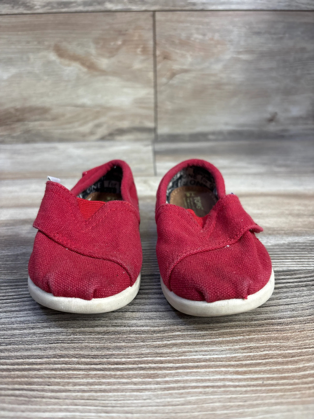 Front view of Toms Tiny Alpargata toddler red canvas slip on shoes showing white rubber soles on wood floor