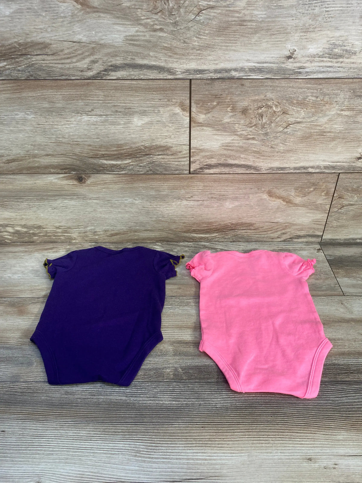 Front view of NFL Team Apparel Baltimore Ravens 2-pack bodysuits in purple and pink for 0-3 months on a wood-look background