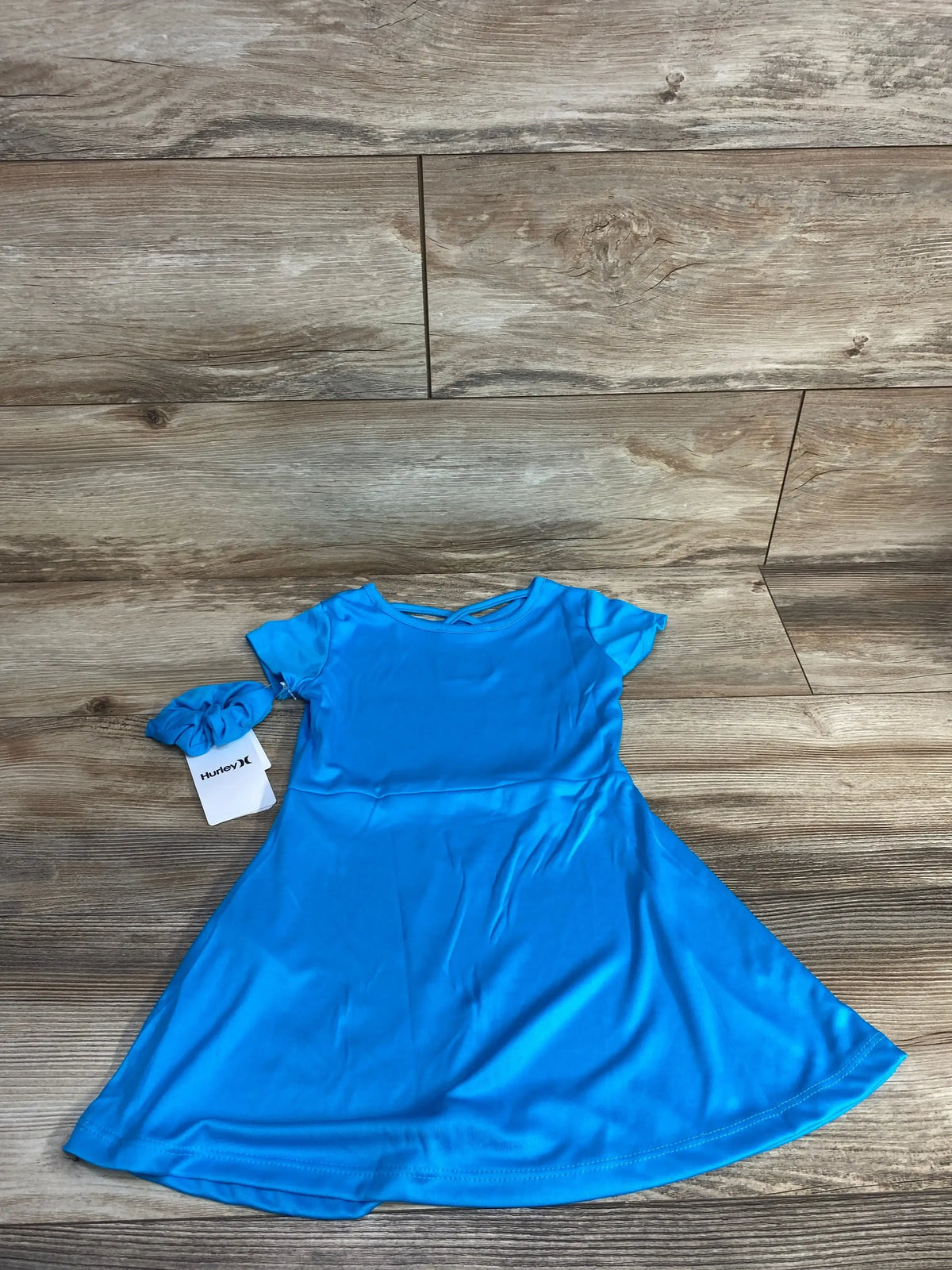 Front flat-lay of blue Hurley toddler dress showcasing short sleeves, flared skirt, and matching scrunchie on wood plank background