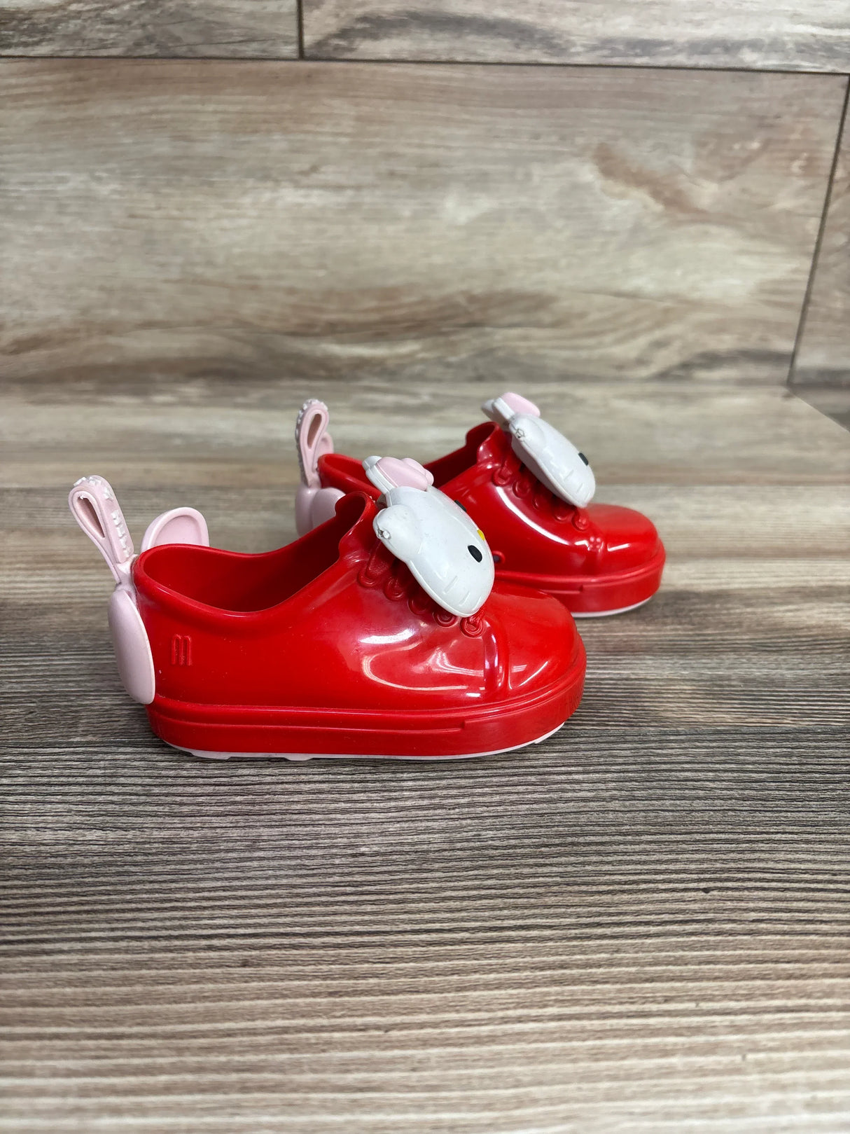 Side view of Mini Melissa Hello Kitty toddler slip-on shoes (size 5C) showing white Hello Kitty face charms and pink heel pull tabs on a wood floor