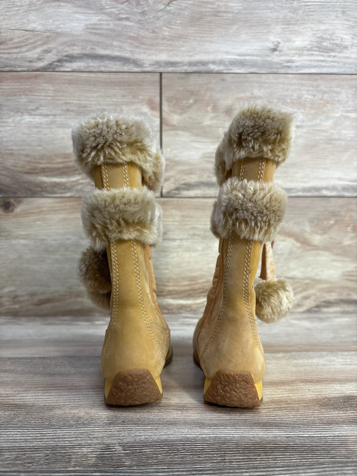 Back view of tan suede winter boots with fur trim and pom-poms on wood floor