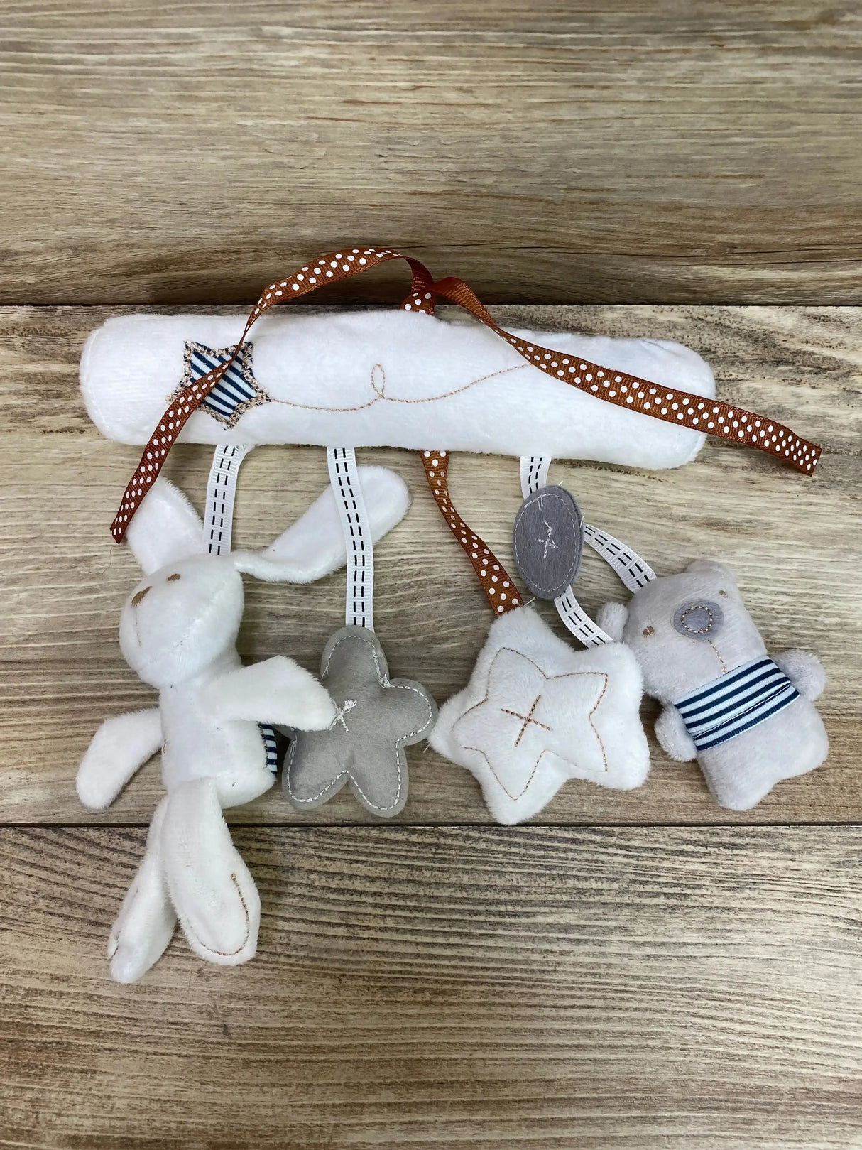 Top-down view of white hanging plush music toy with rabbit, bear, two star shapes, striped fabric accents, and polka-dot attachment ribbon on wood background