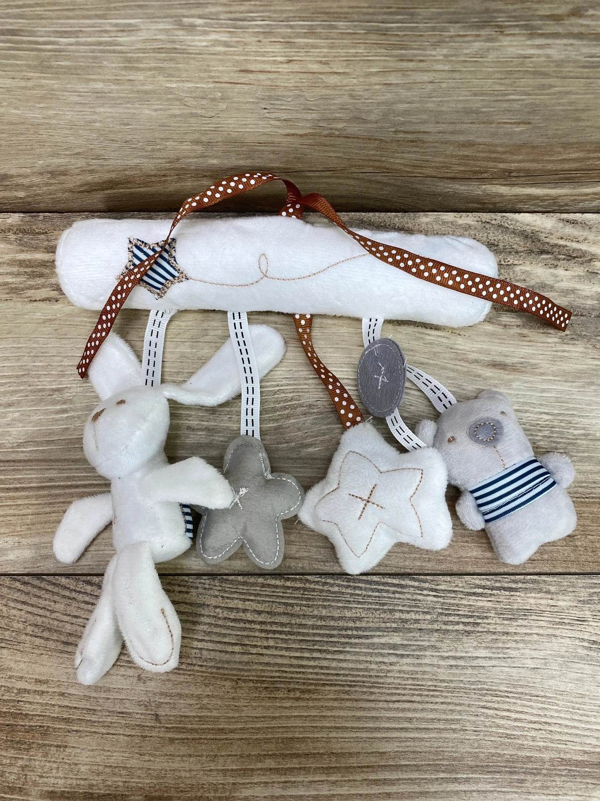 Top-down view of white hanging plush music toy with rabbit, bear, two star shapes, striped fabric accents, and polka-dot attachment ribbon on wood background