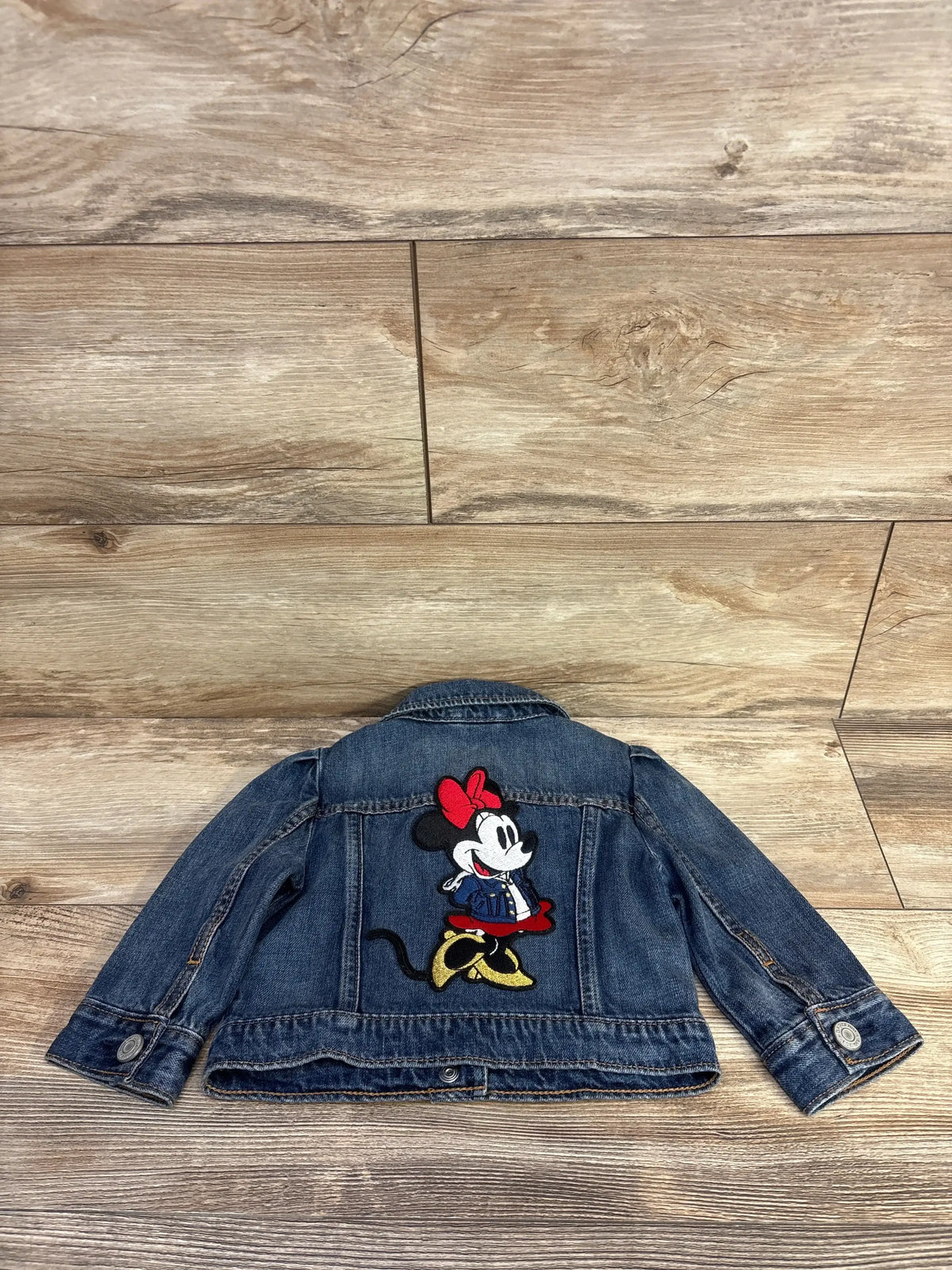 Back view of Baby Gap x Disney baby denim jacket showcasing large embroidered Minnie Mouse patch, button cuffs on wood floor