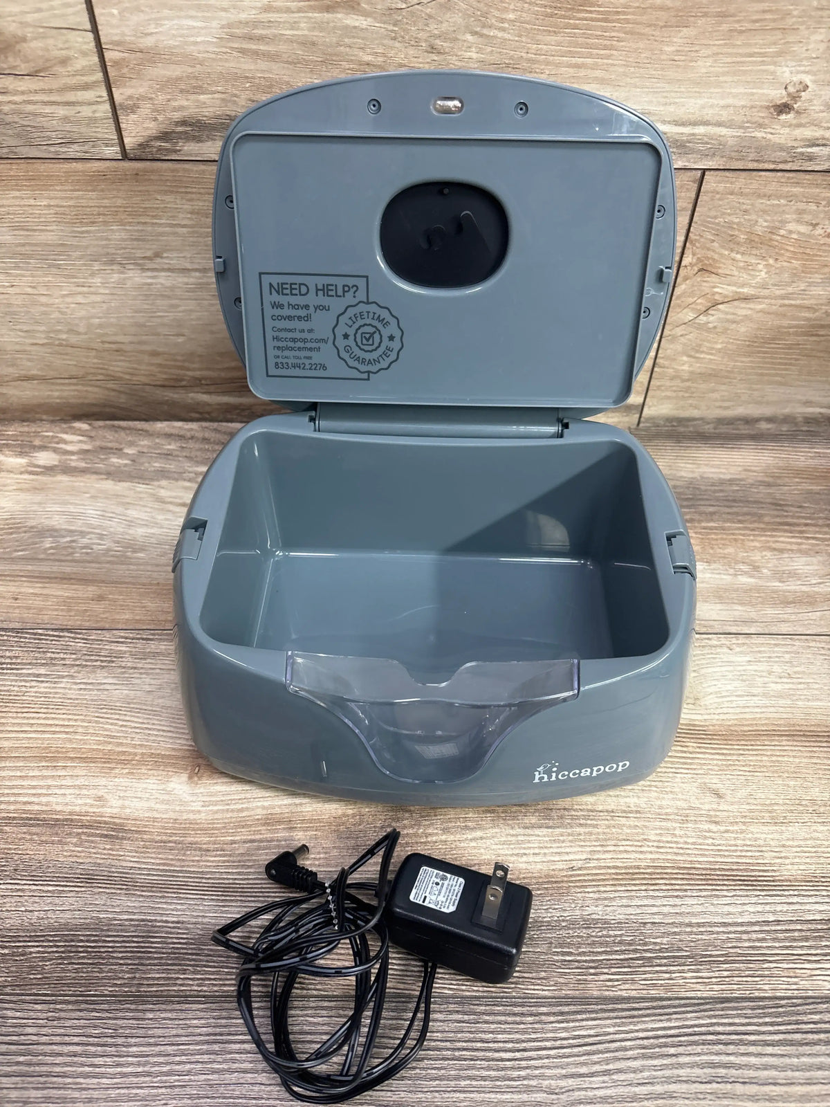 Open view of Hiccapop Ultra Wipe Warmer in grey showcasing hinged lid, empty basin, front cover, and power adapter on a wood floor