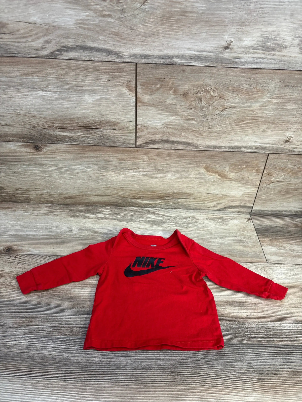 Front view of red Nike long-sleeve baby shirt, size 9 months, showing black Nike wordmark and swoosh on light wood floor