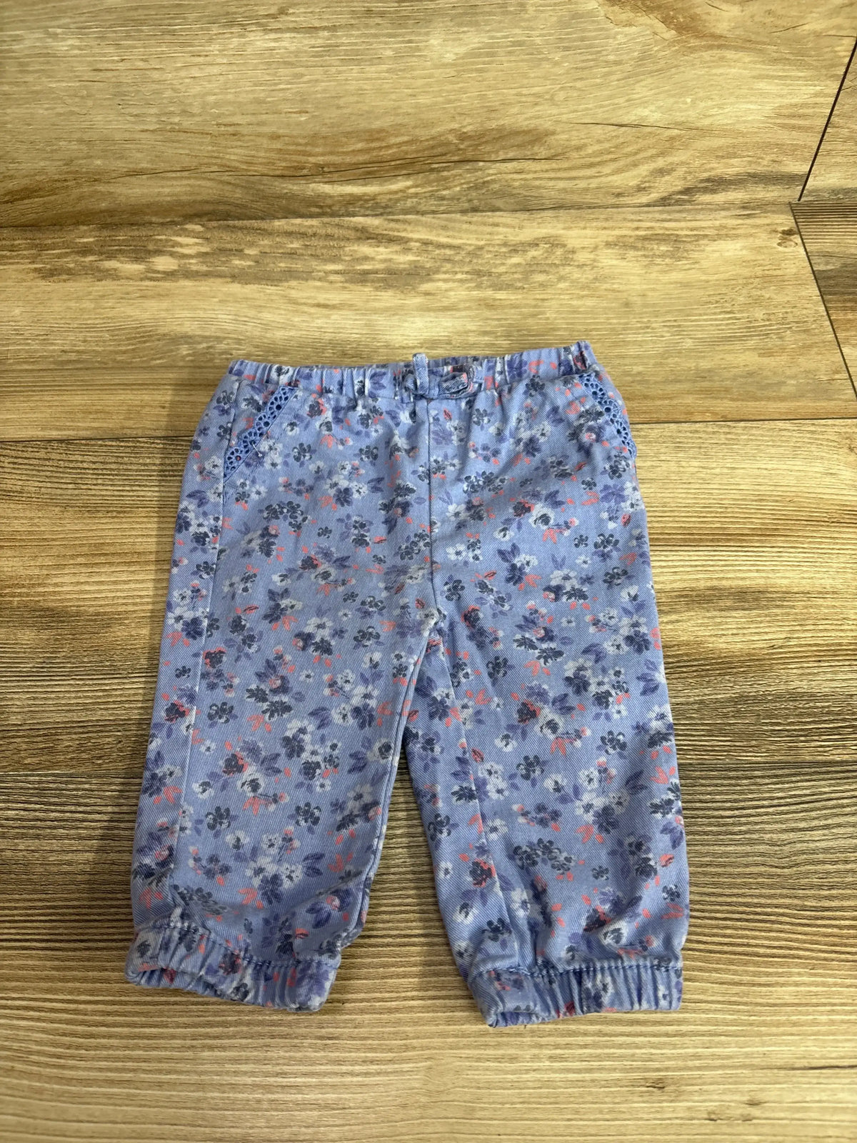 Front view of blue Oshkosh floral print pull-on twill pants showcasing lace pocket detail and elastic ankles on a wood surface