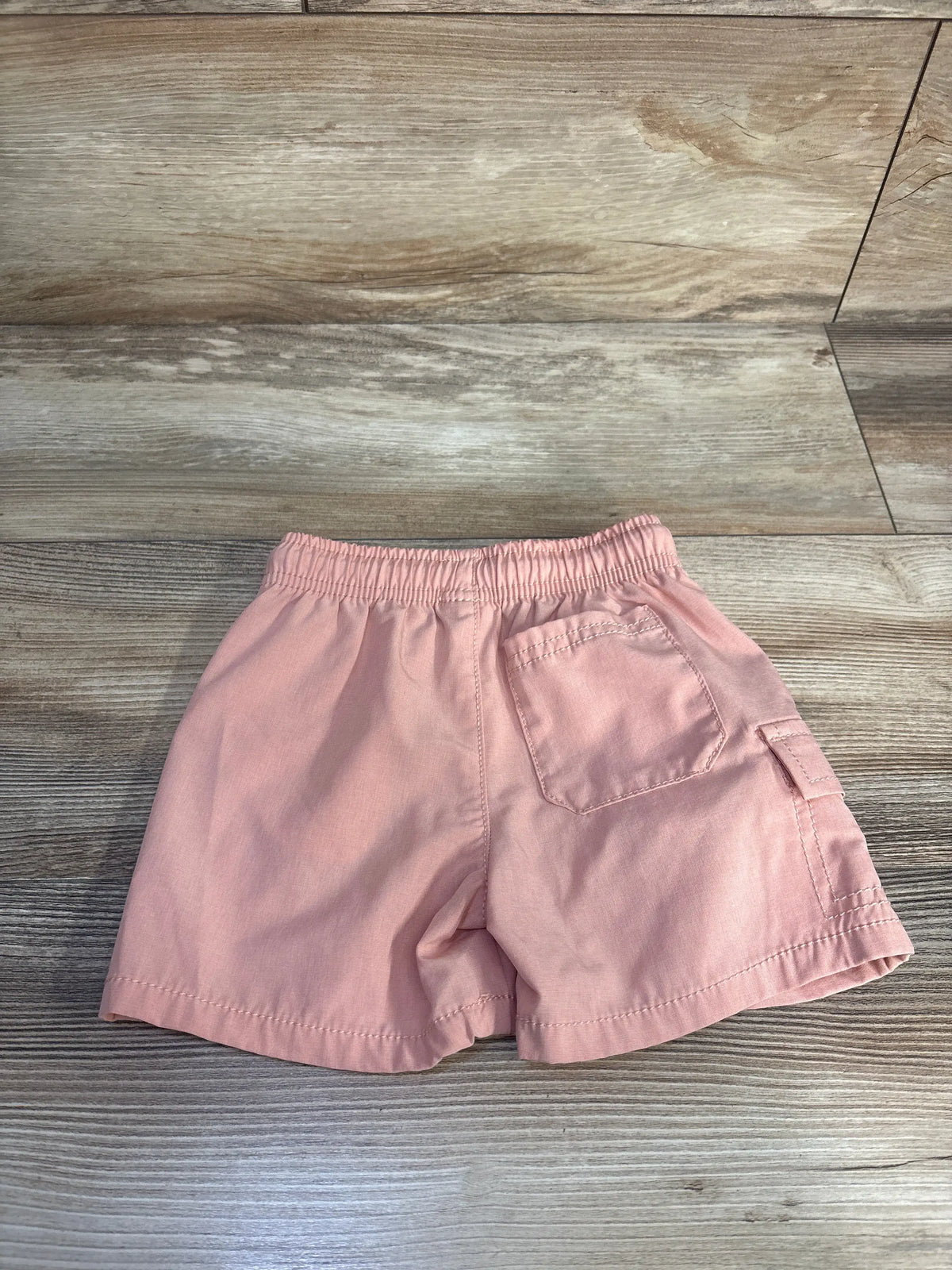Rear view of Oshkosh orange board shorts size 18 months with elastic waistband and back and side pockets on wood-look flooring