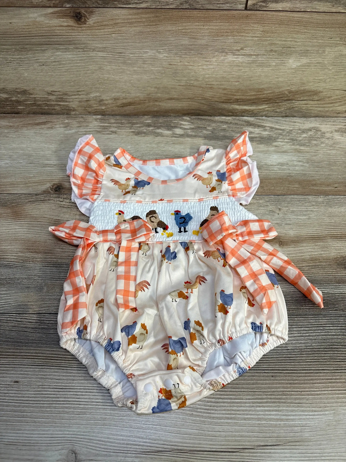Top-down view of ivory bubble romper with smocked chest, embroidered roosters, orange gingham bows and flutter sleeves on wood, size 12-18 months
