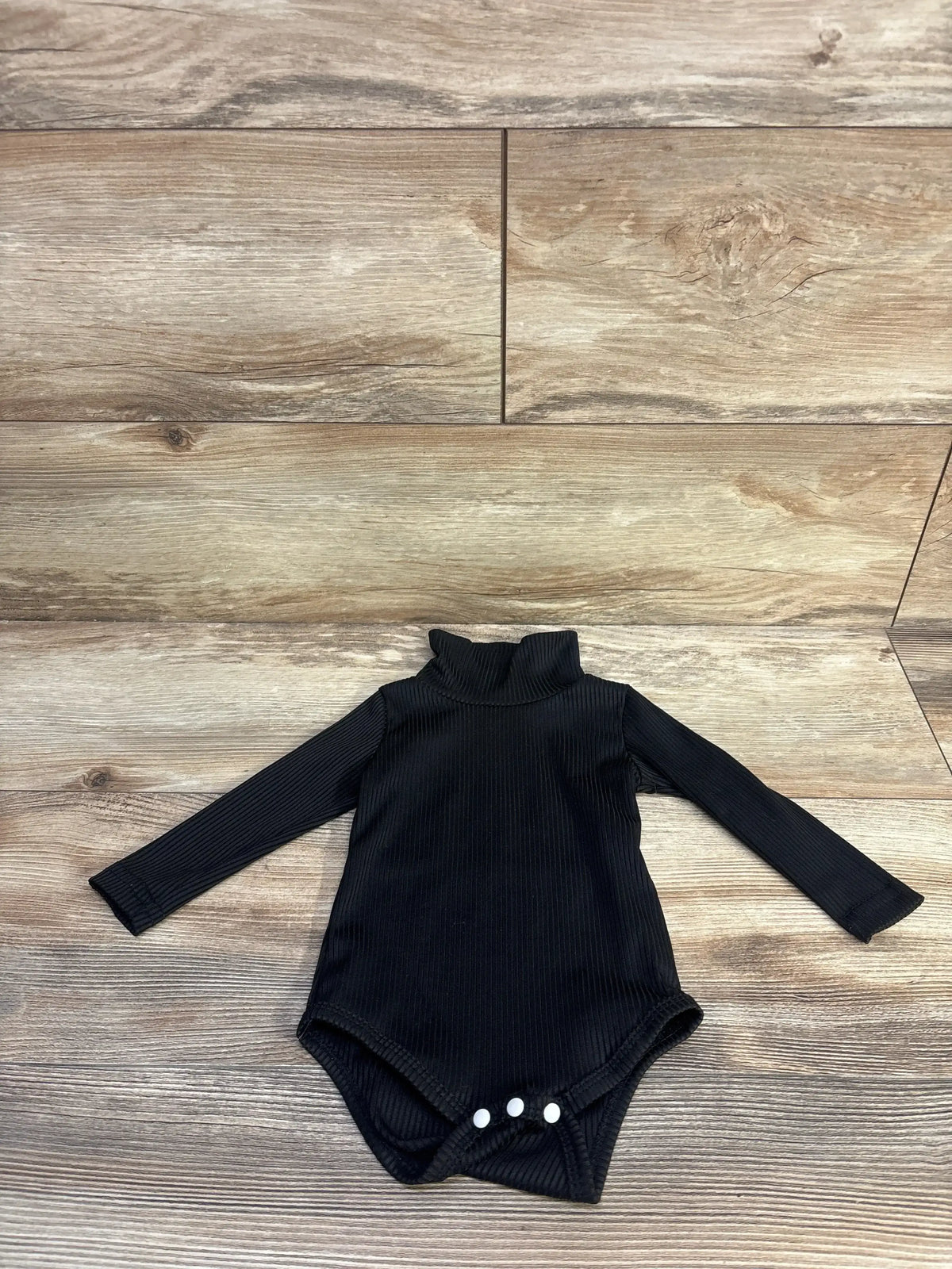 Flat lay of black ribbed turtleneck baby bodysuit showing three snap crotch on wood background