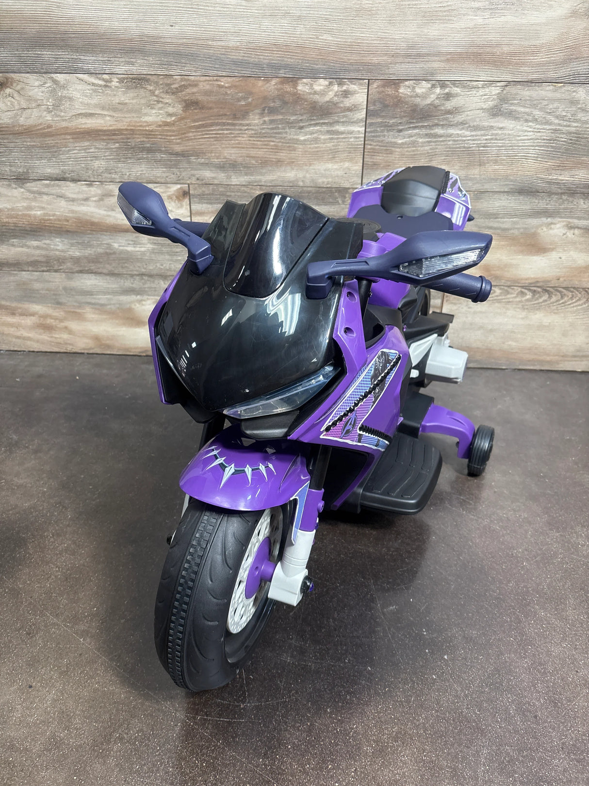 Front three-quarter view of Marvel Black Panther ride-on motorcycle showcasing purple and black body, windshield, and training wheels on wood backdrop