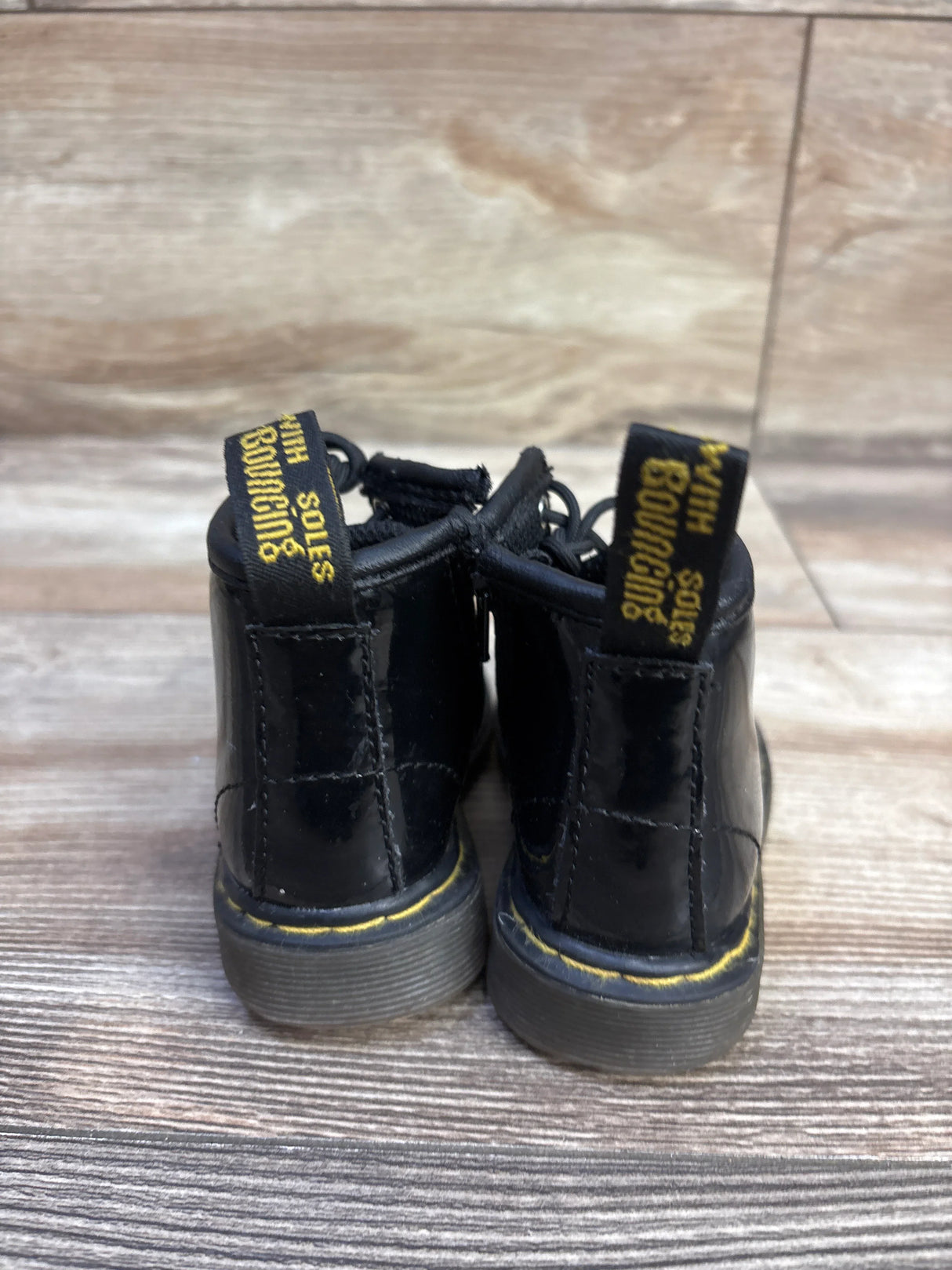 Rear view of toddler Dr. Martens black patent leather ankle boots showcasing yellow welt stitching and heel pull tabs on wood floor