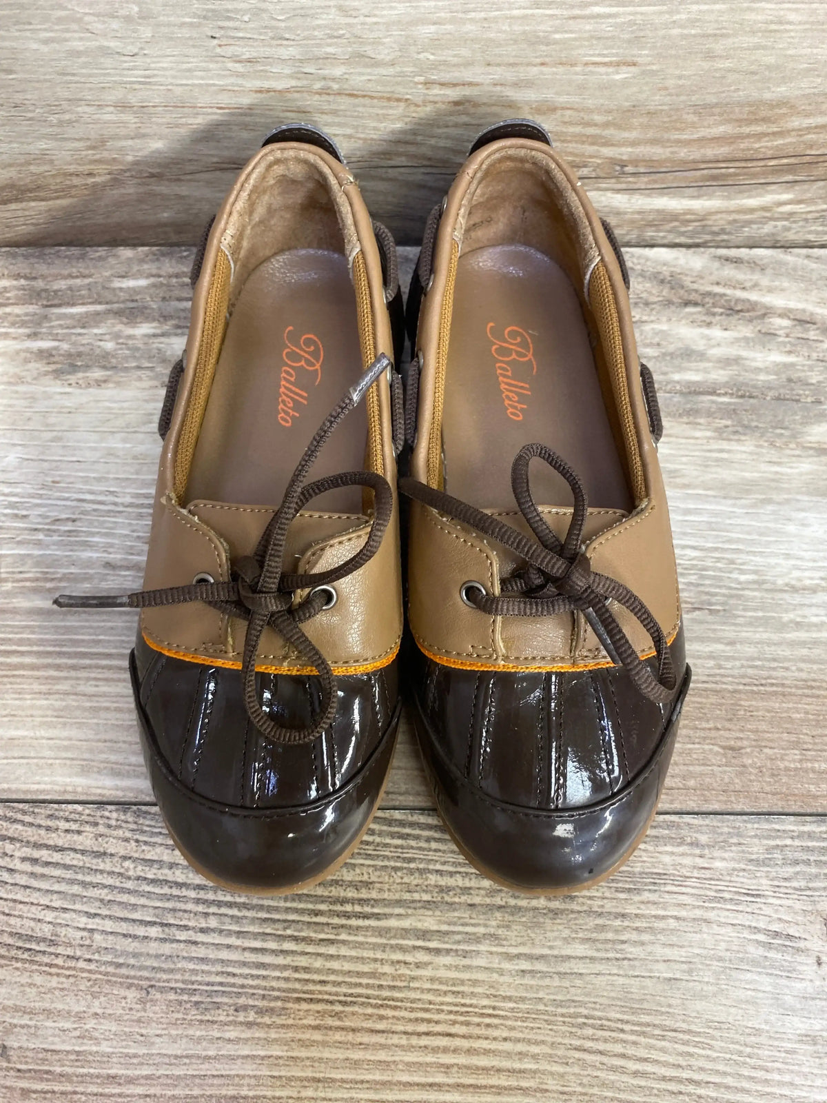 Top-down view of toddler Balleto boat shoes in tan and dark brown showcasing glossy duck-toe and tied laces on a wood plank background