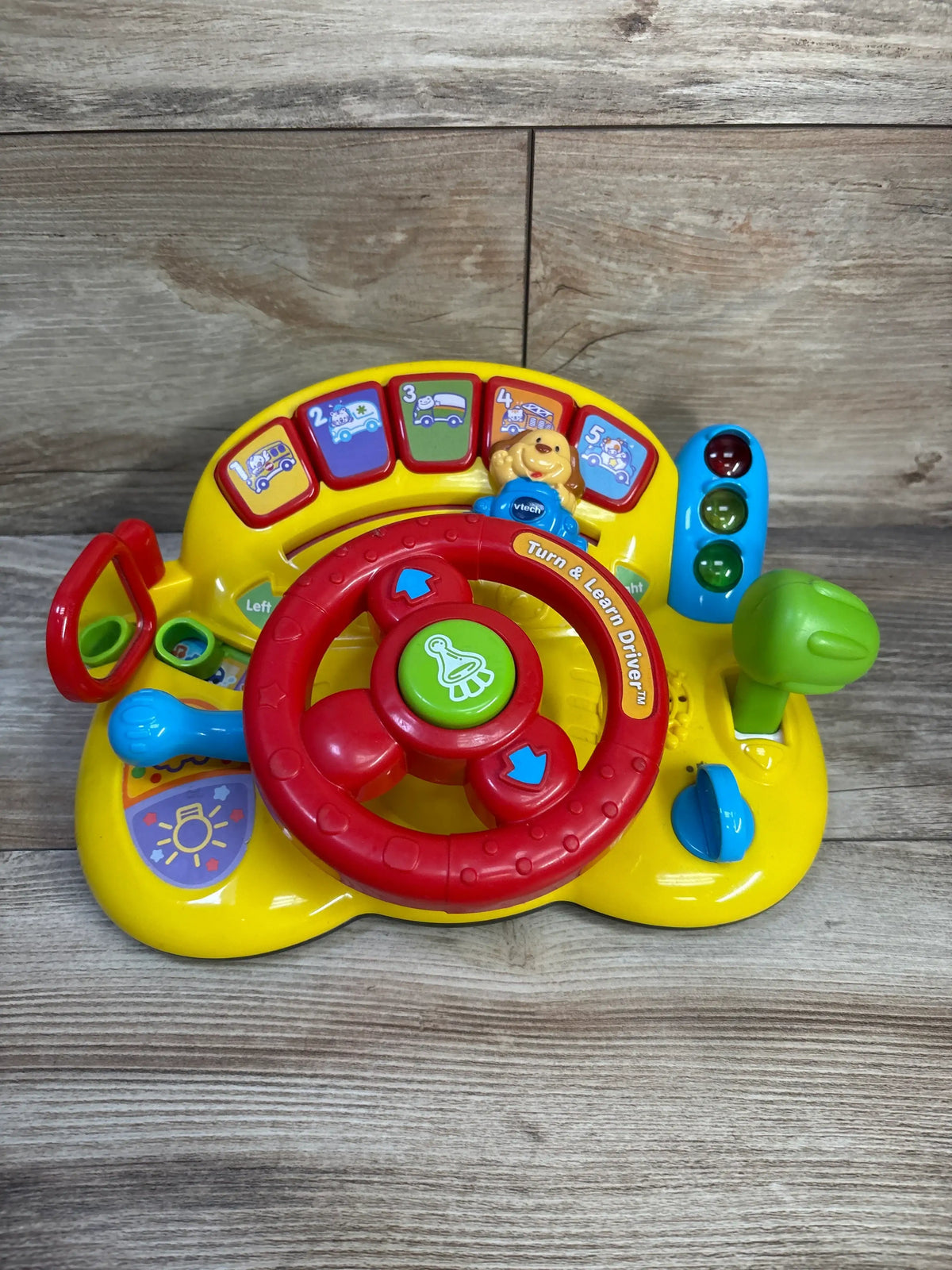 Front view of VTech Turn & Learn Driver showcasing red steering wheel, numbered buttons, gear shifter, and small traffic light on wooden tabletop background