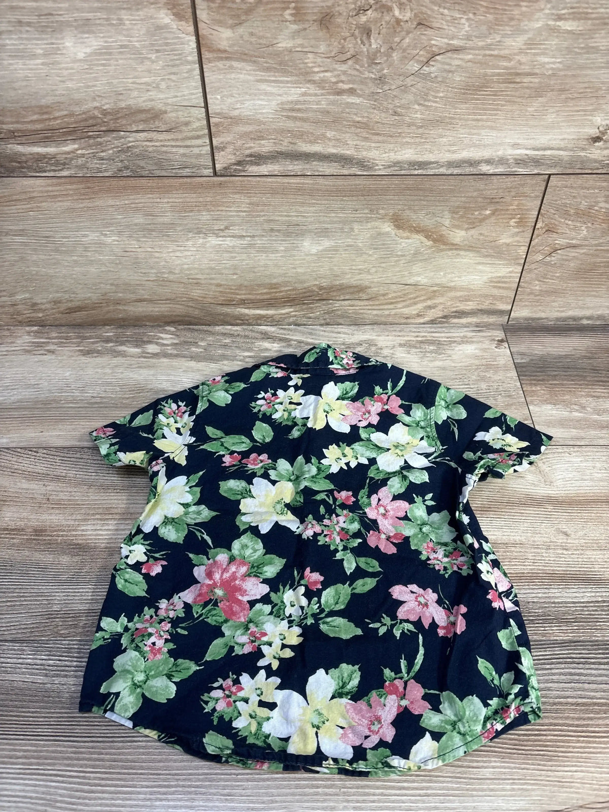 Back view of navy floral short-sleeve toddler button-up shirt showing all-over pink, white, and yellow flower print and curved hem on wood floor background