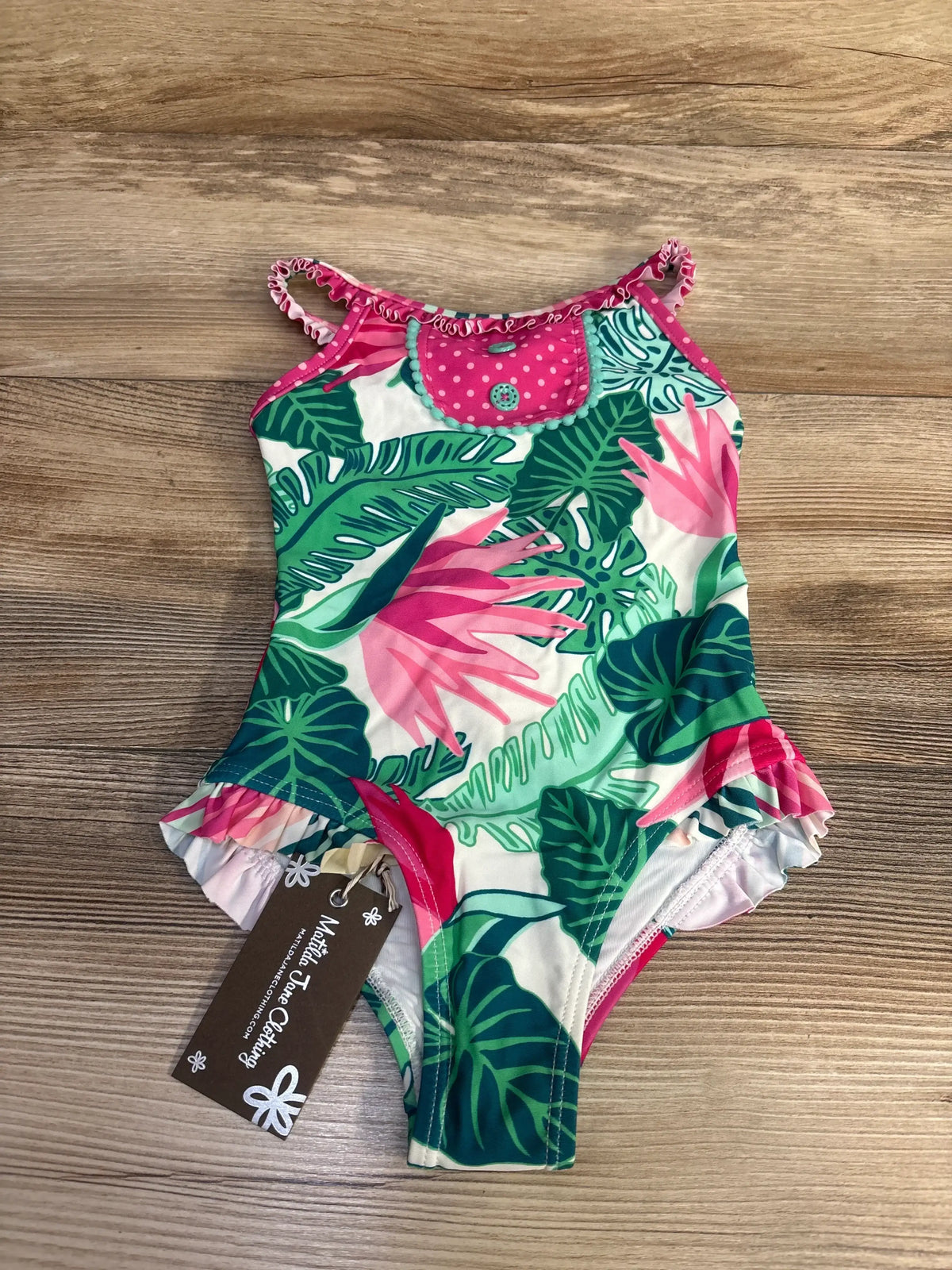 Front view of NWT Matilda Jane tropical ruffle swimsuit with pink and green palm leaf print and pink polka dot bib on wood plank floor