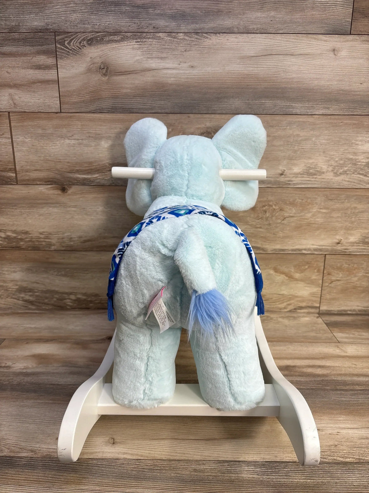 Rear view of pale blue plush elephant nursery rocker showing patterned blue saddle and white wooden rocker base against wood plank background