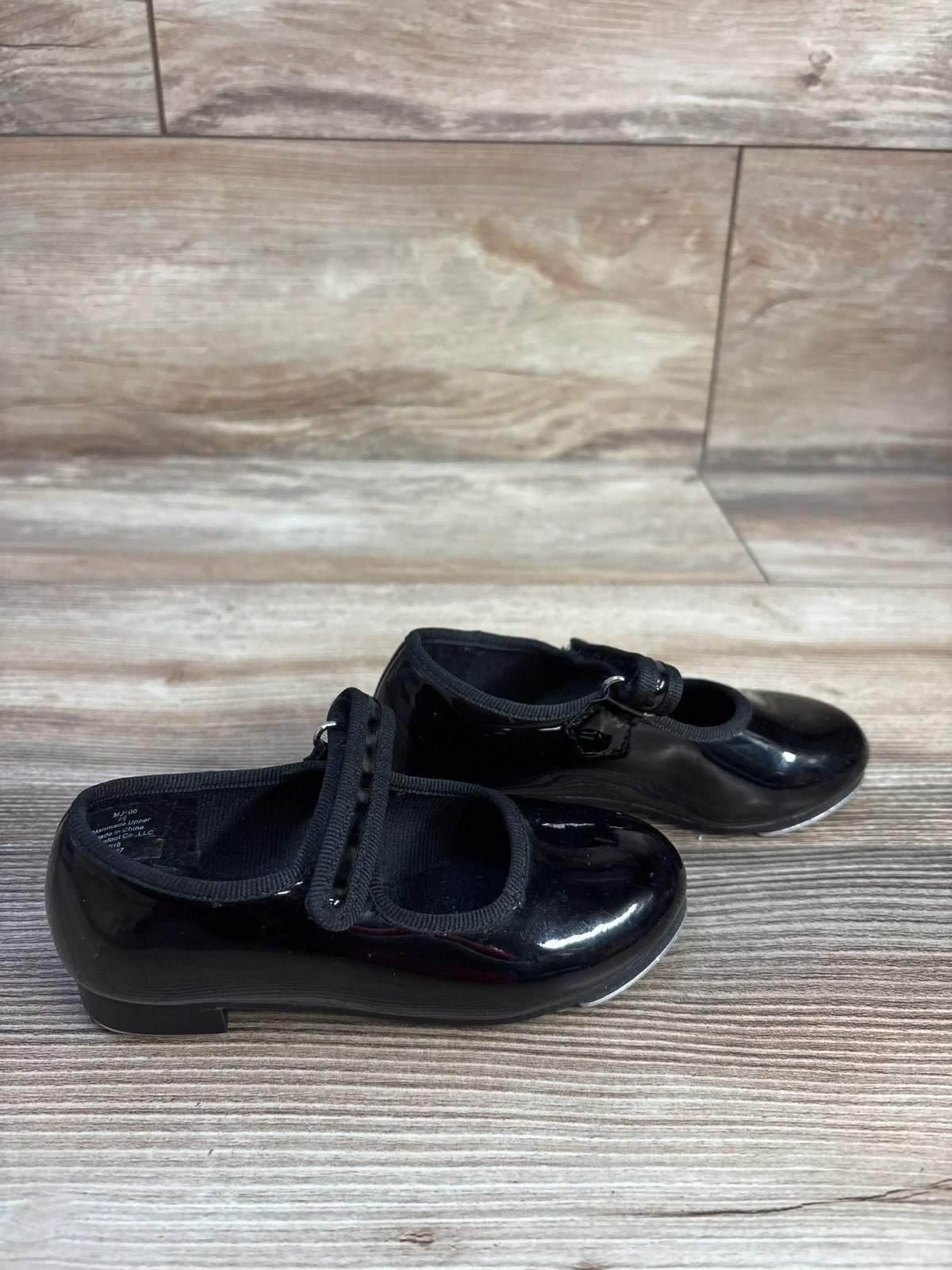Three-quarter view of black tap shoes in size 7.5c showcasing Mary Jane straps with a wood-look floor background