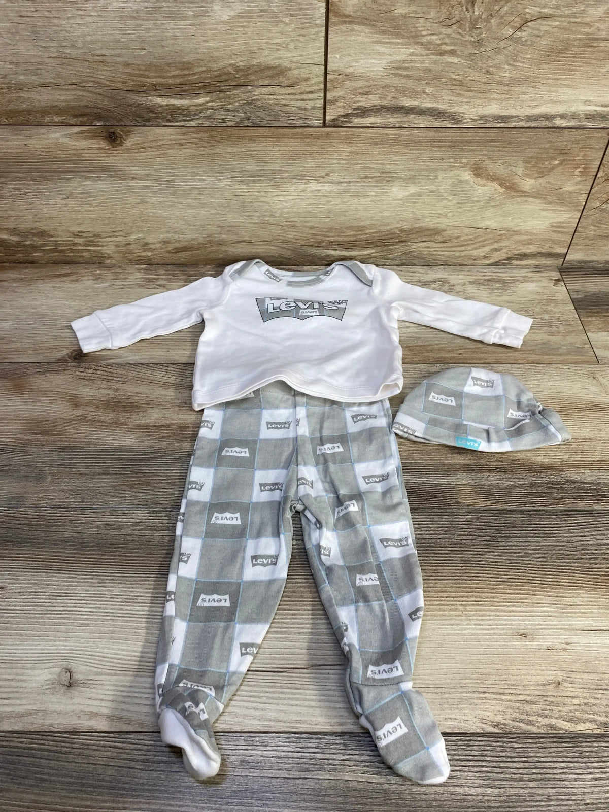 Flat lay of baby Levi’s 3-piece set with white logo top, gray checkered footed pants and matching hat on wood floor