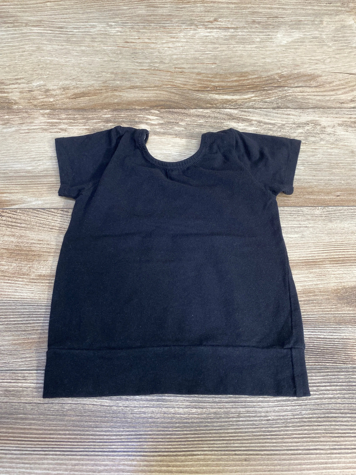 Flat-lay of black short-sleeve toddler shirt showing crew neckline, short sleeves, and wide hem on light wood background