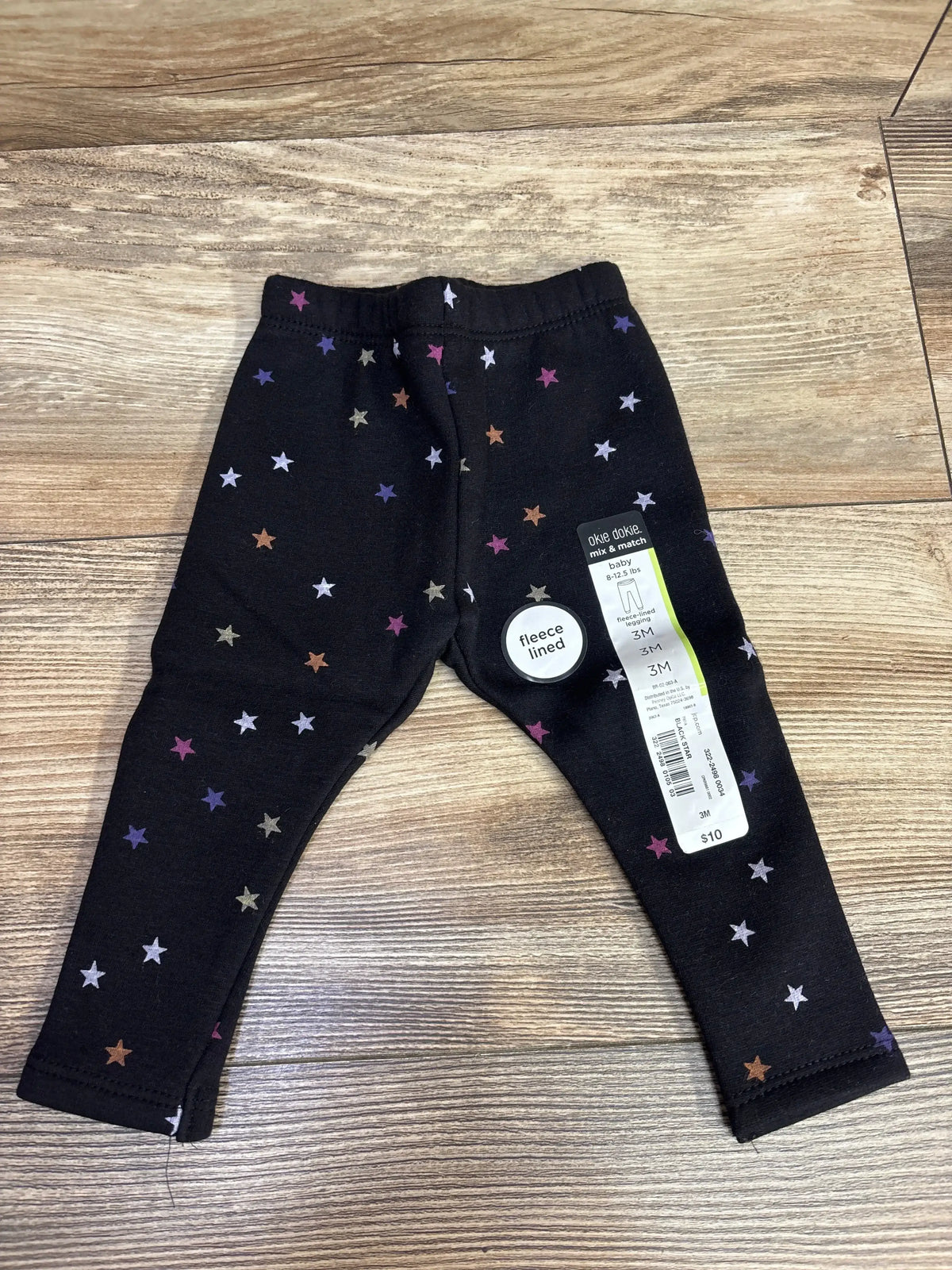 Flat lay of black fleece-lined baby leggings with multicolor star print showing elastic waistband on wooden floor, size 3M