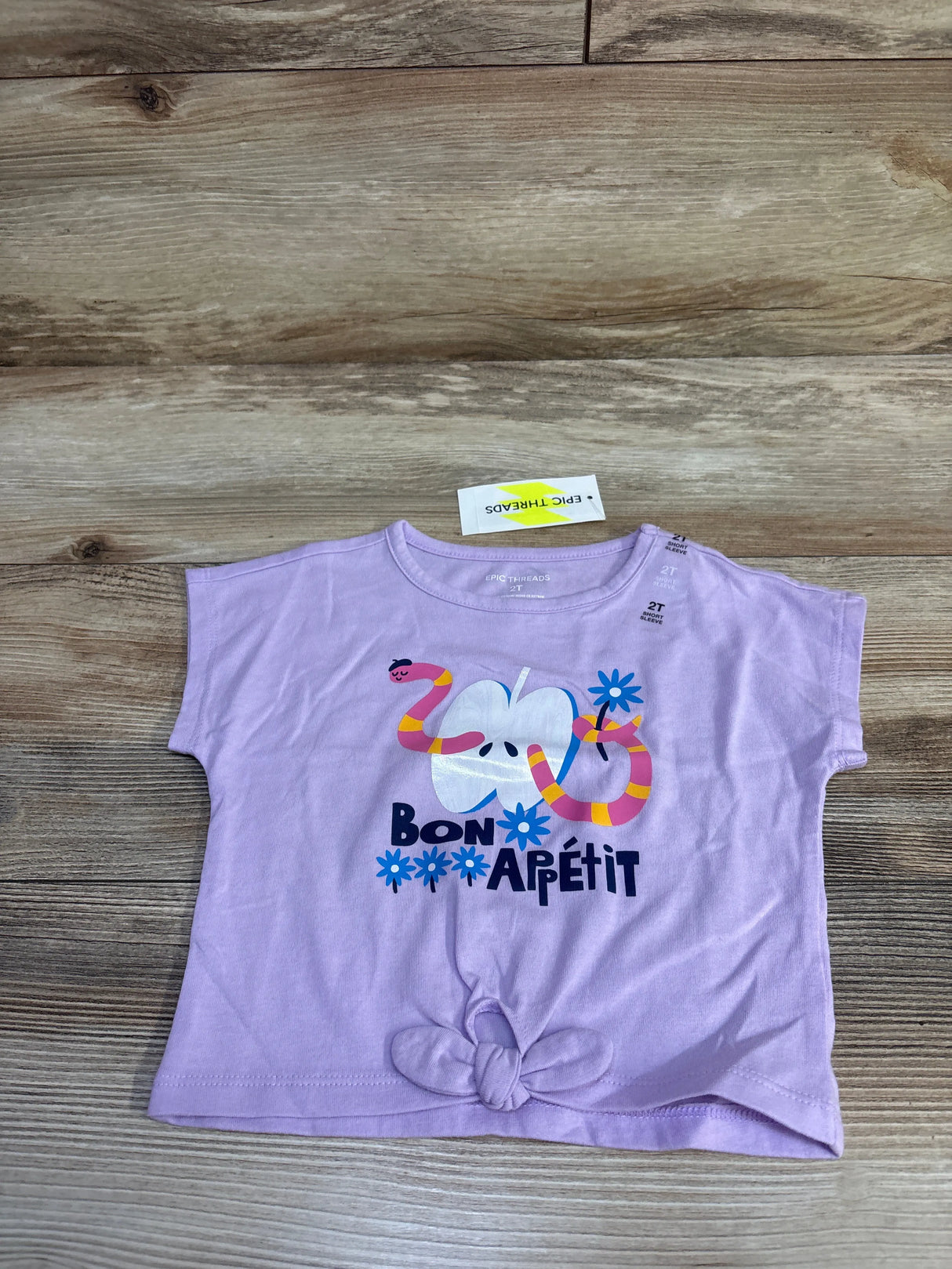 Front flat lay of Epic Threads toddler t-shirt in lilac showing Bon Apétit apple and snake graphic with knotted hem on wood
