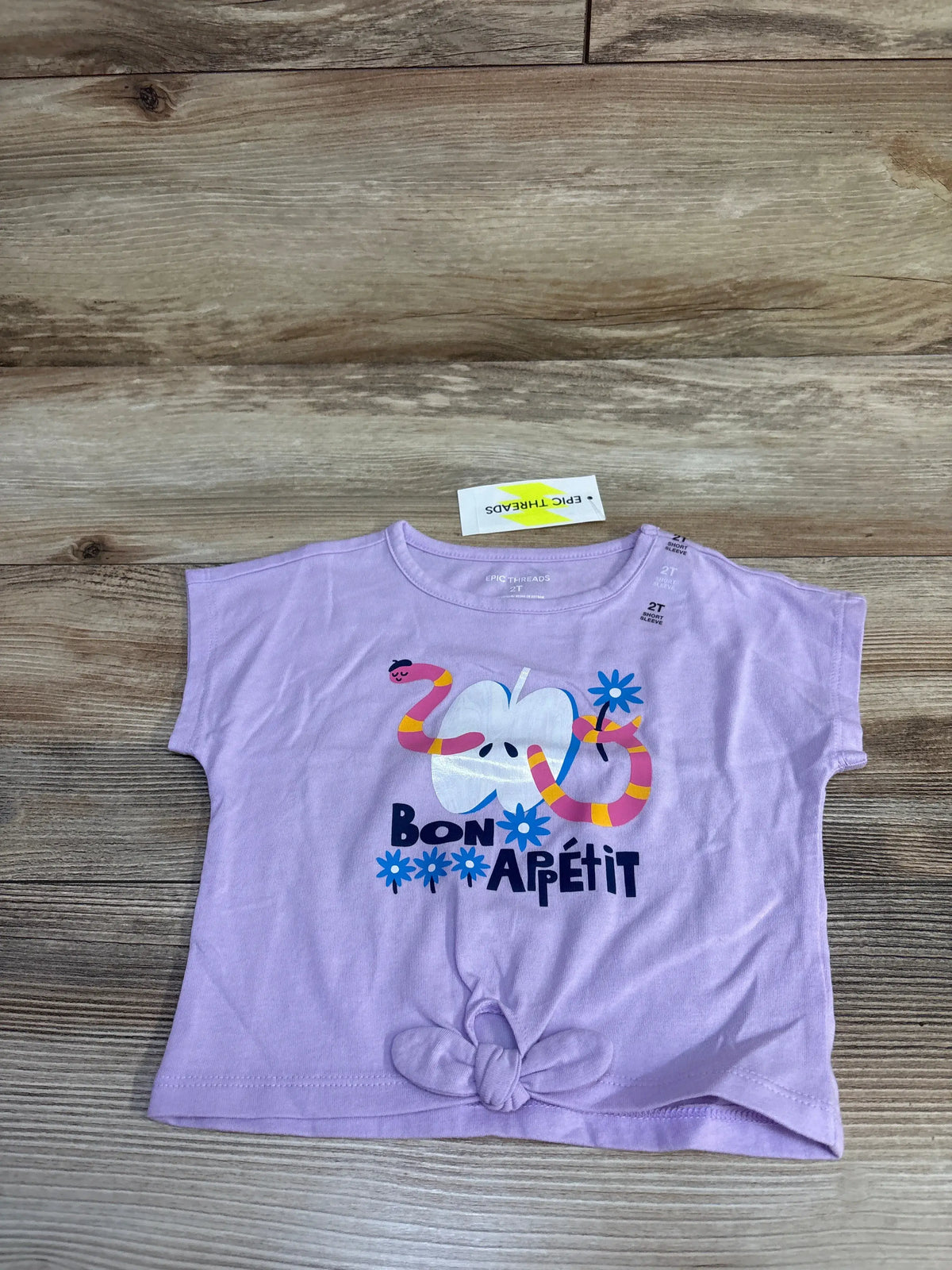 Front flat lay of Epic Threads toddler t-shirt in lilac showing Bon Apétit apple and snake graphic with knotted hem on wood
