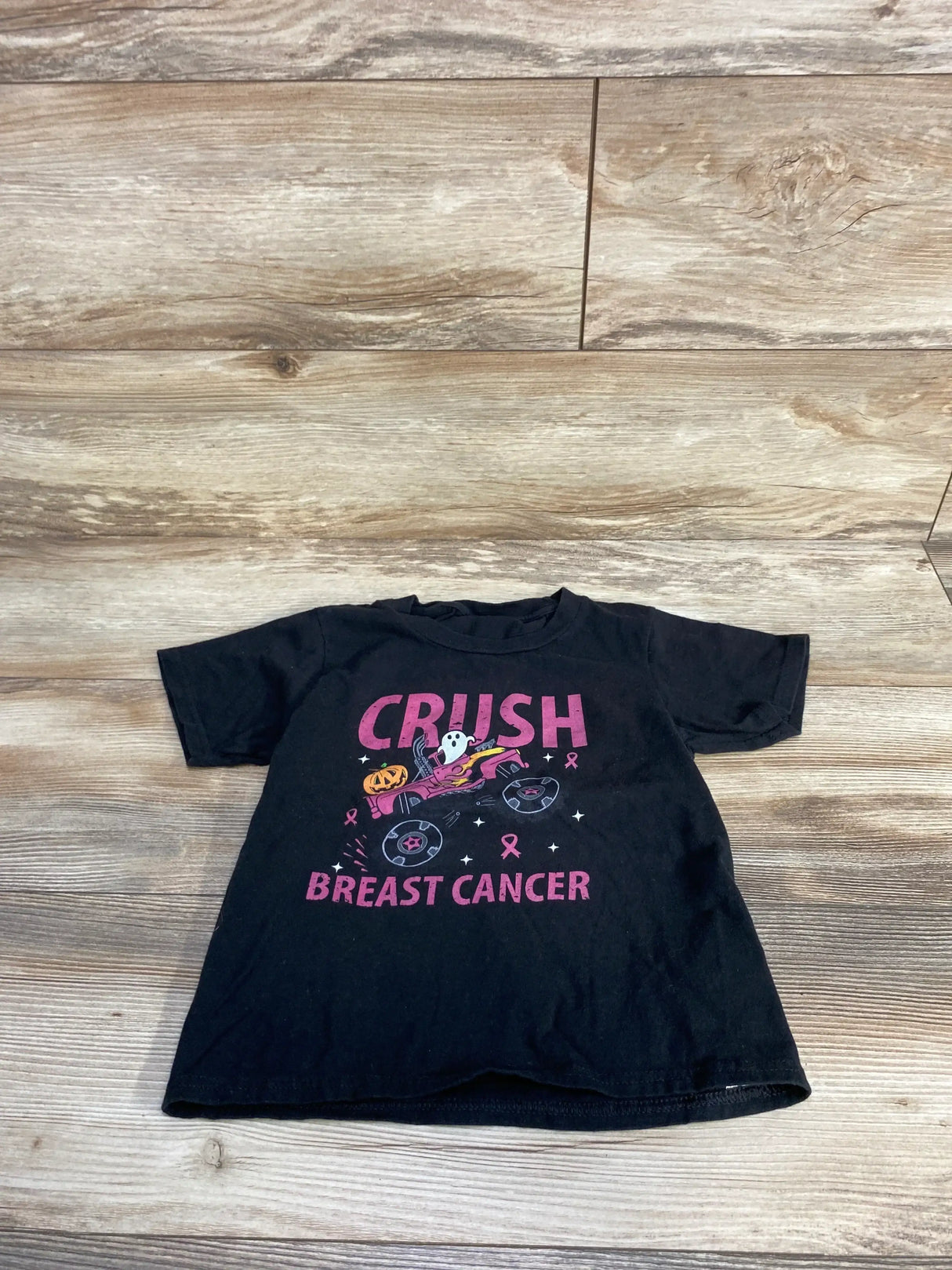 Black kids t-shirt with 'Crush Breast Cancer' pink text, ghost, pumpkin, and ribbons on wood floor