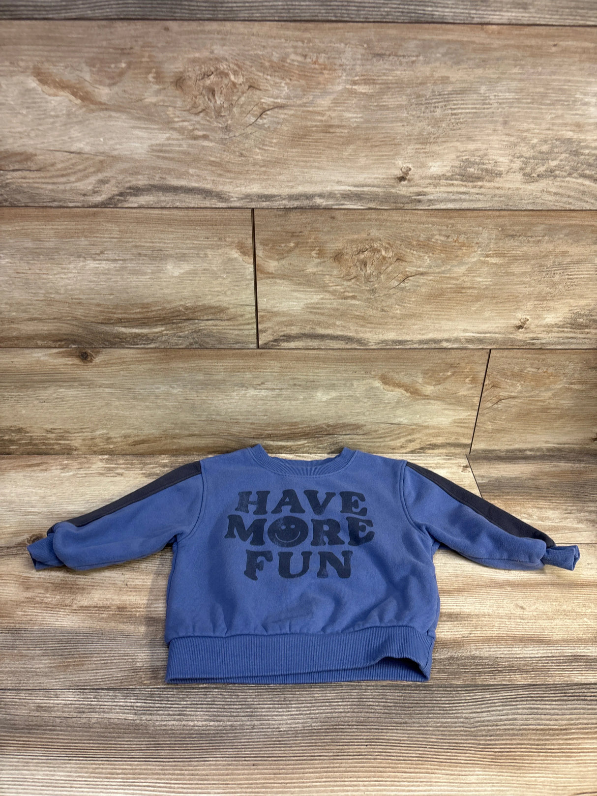 Front flat-lay of blue toddler pullover sweatshirt showing bold HAVE MORE FUN black print and contrasting black shoulder stripes on wood background