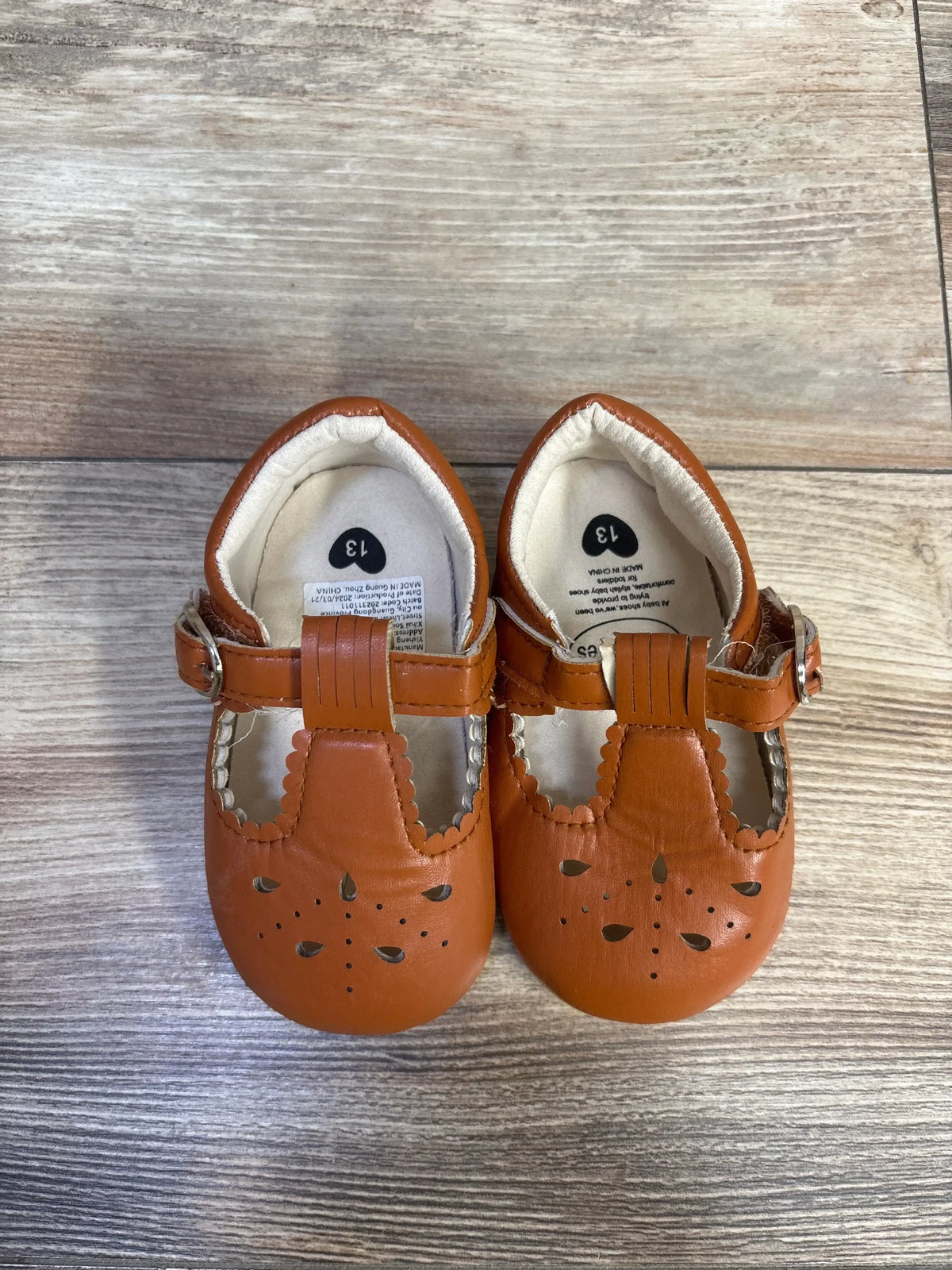 Top-down view of chestnut Sienna toddler sandals showcasing scalloped edges, floral cutouts, and hook and loop straps on wood