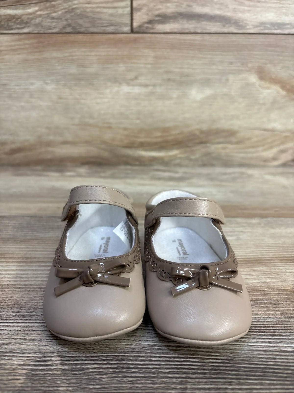 Front view of toddler Mary Jane soft-sole flats showcasing taupe faux-leather upper, glossy bow and scalloped trim on wood background