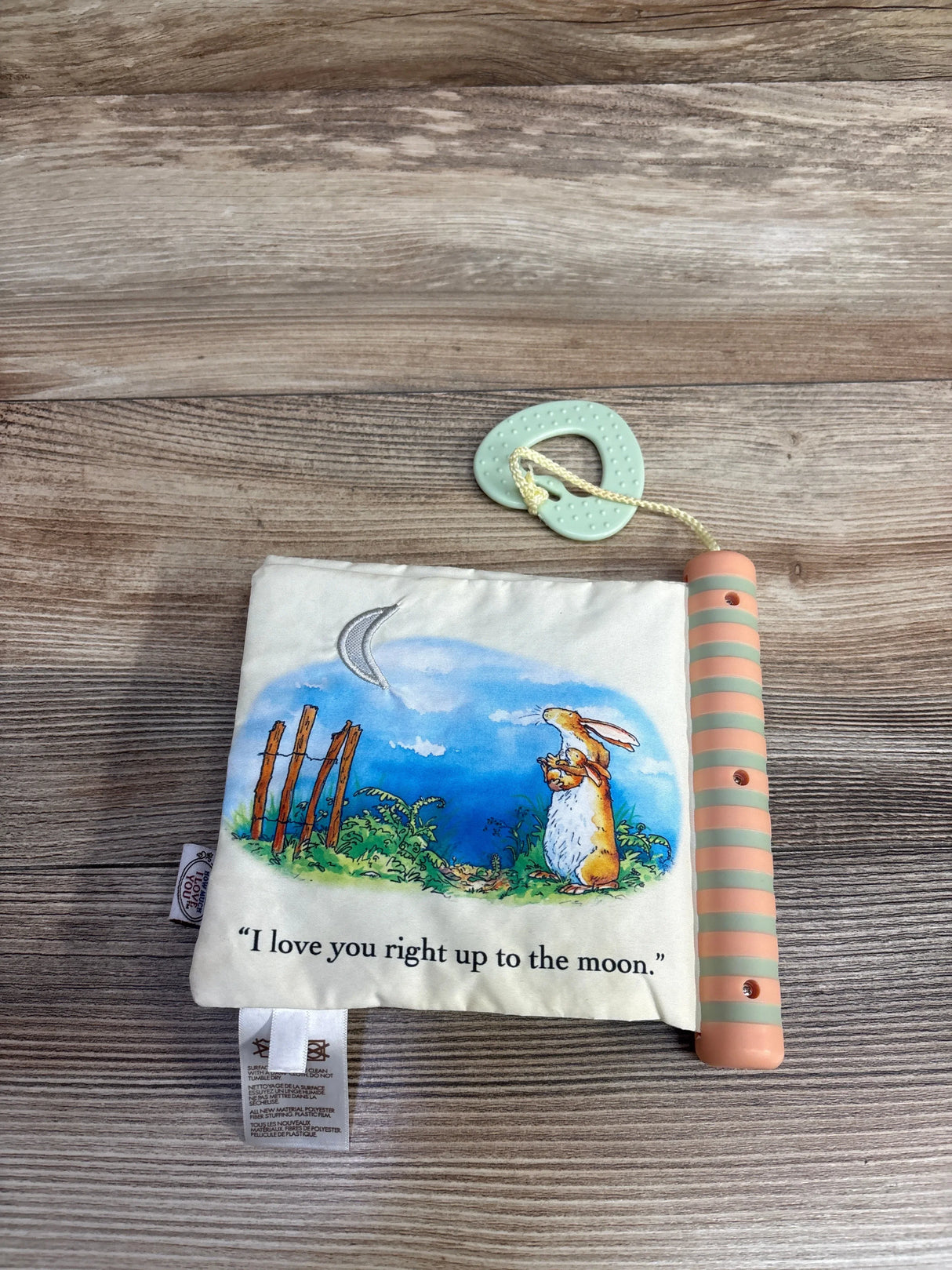 Soft baby cloth book with bunny illustration, teether, and quote, on wood surface.