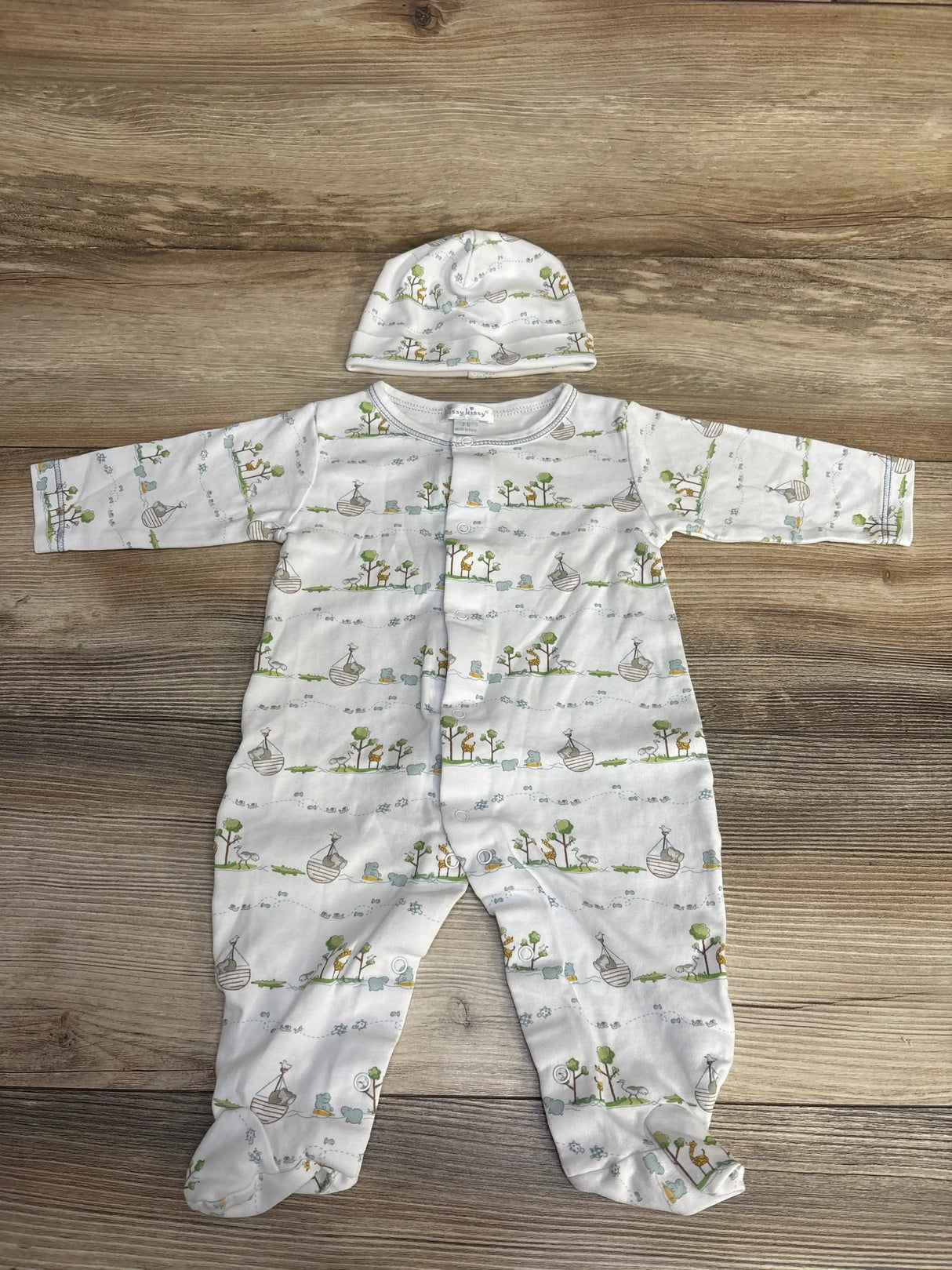 Flat lay of Kissy Kissy white footed baby romper and matching hat showing front snaps and Noah’s Ark print on wood background