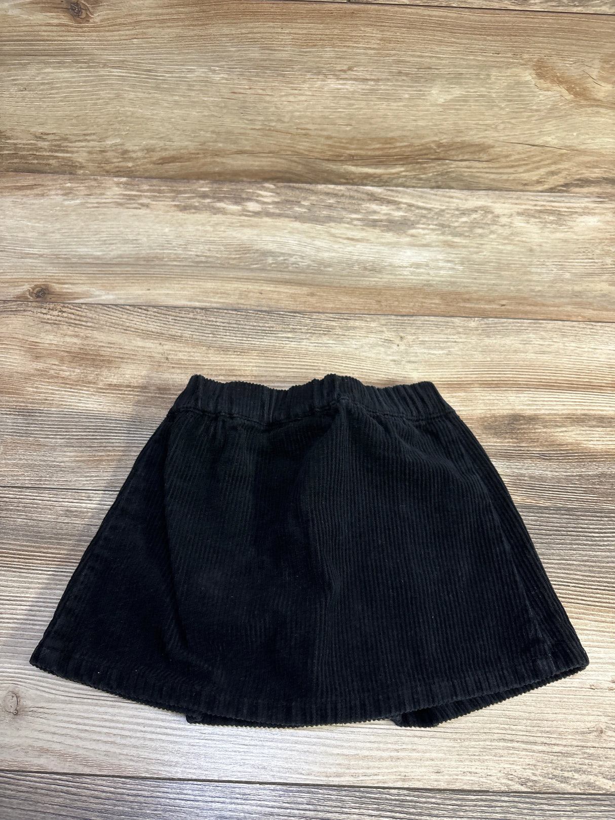 Top view of Art Class corduroy skirt, black, showing elastic waistband and vertical ribs on wood background