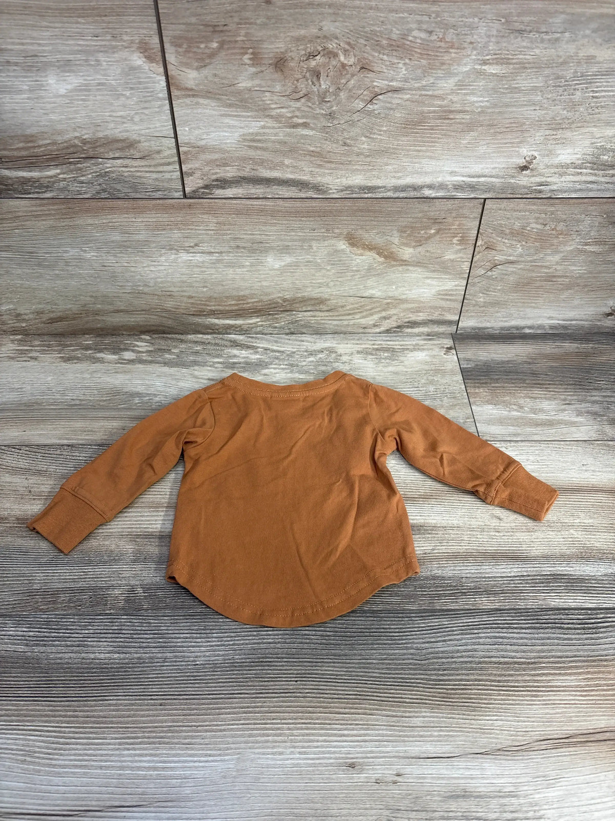 Back view of Little Bipsy long-sleeve newborn tee in rust orange showing curved hem and cuffed sleeves on wood background