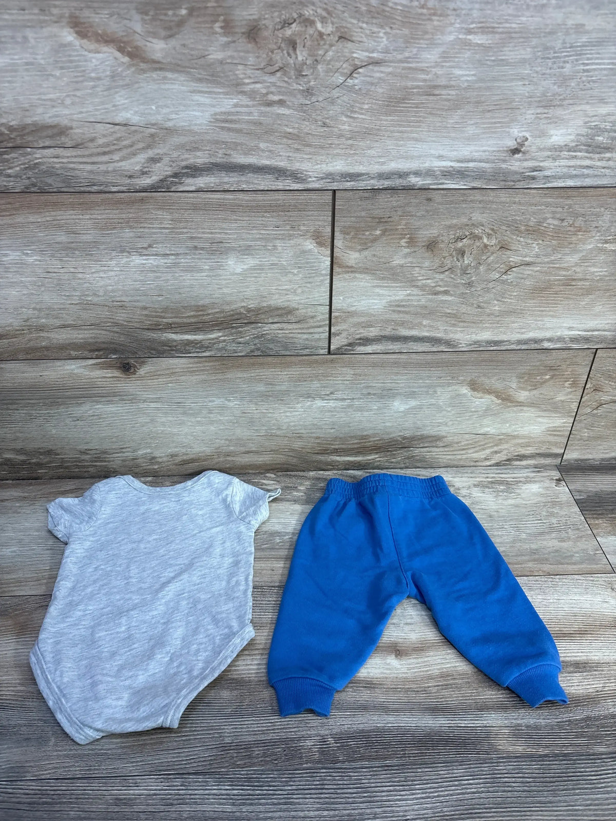 Flat lay of Nike 2-piece grey short-sleeve bodysuit and blue elastic-waist pants size 3M on wood plank background