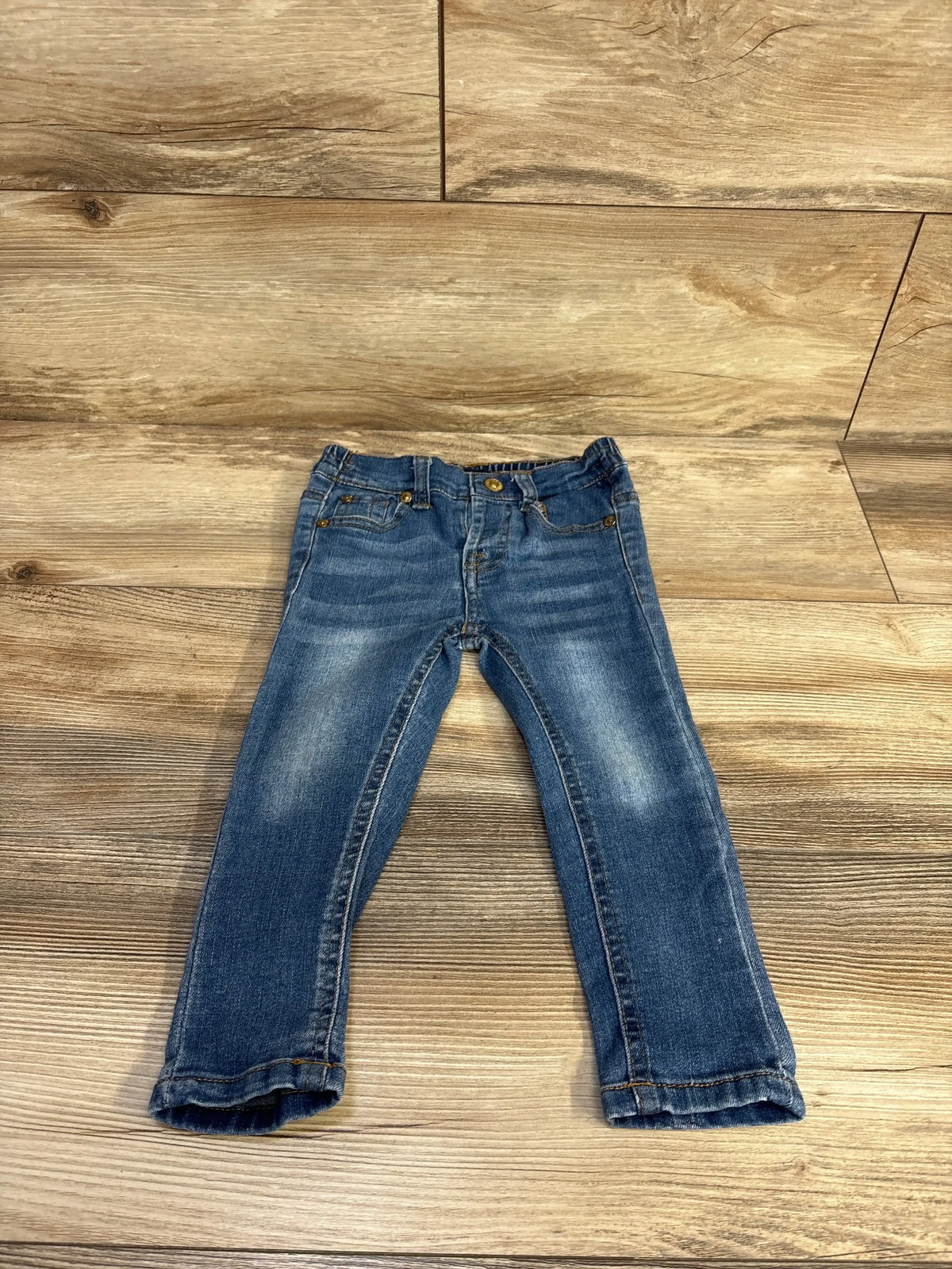 Flat lay of 7 For All Mankind toddler jeans, size 24 months, showing faded knees and rolled cuffs on light wood floor