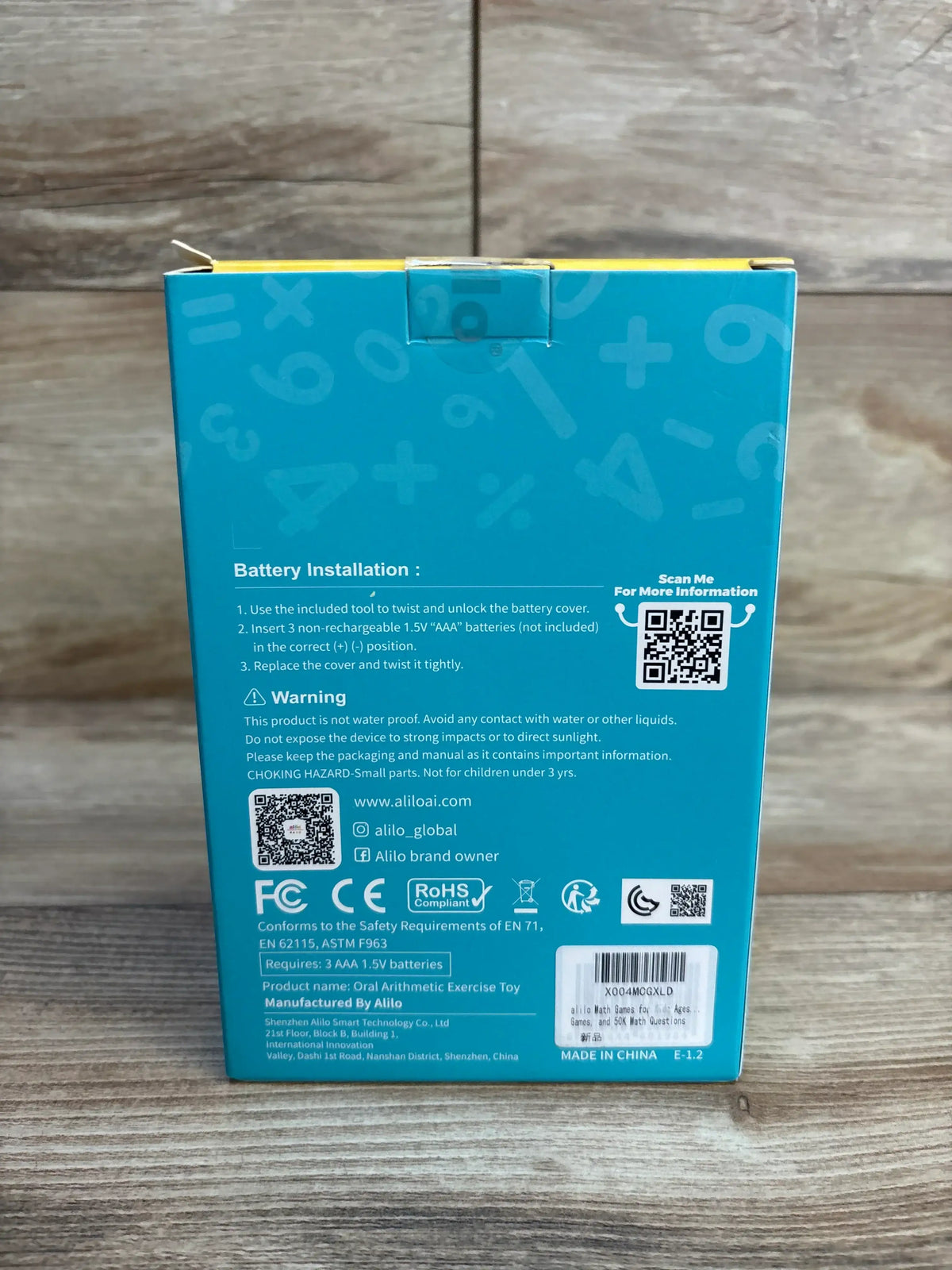 Back view of Alilo Math Magic portable math game box showing battery installation steps, safety warnings, certifications, and barcode on a wood surface