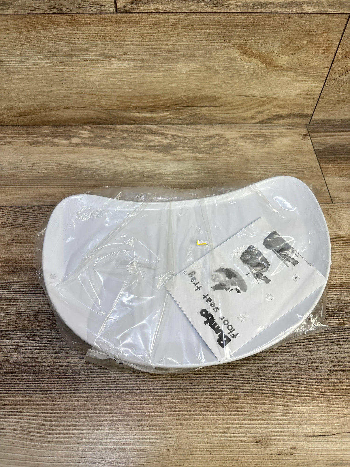Overhead view of white Bumbo seat tray attachment in clear packaging with instruction sheet on wood floor