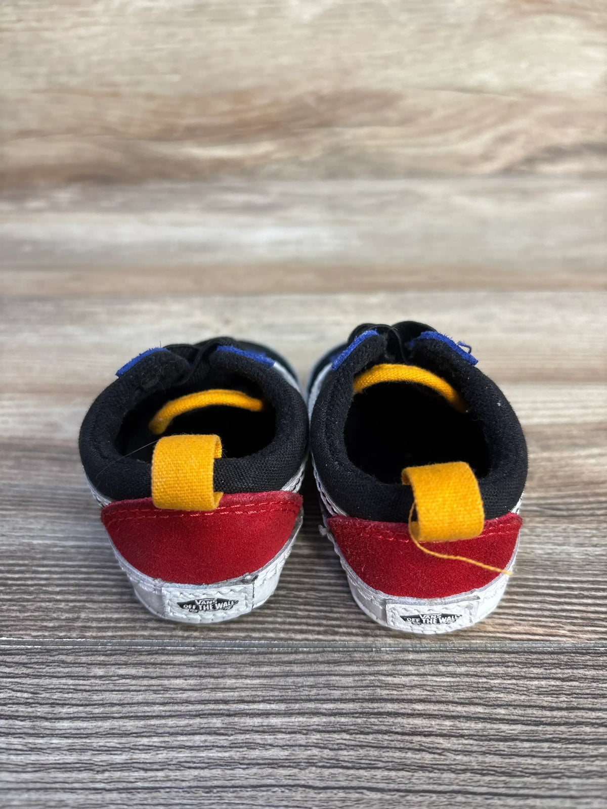 Rear view of black Vans Old Skool crib shoes size 2C with heel pull tabs and waffle sole print on wood background