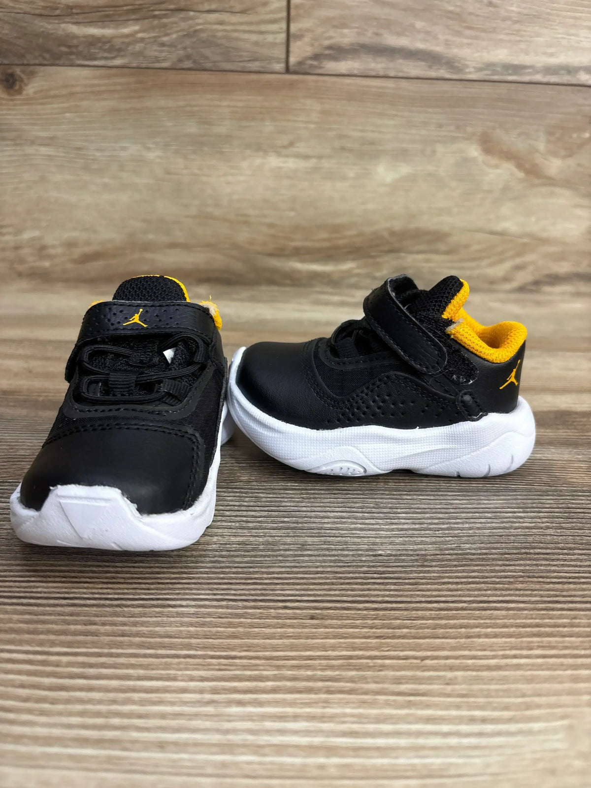 Front and side view of Air Jordan 11 CMFT Low TD Black Taxi toddler sneakers with yellow lining and white soles on wood floor