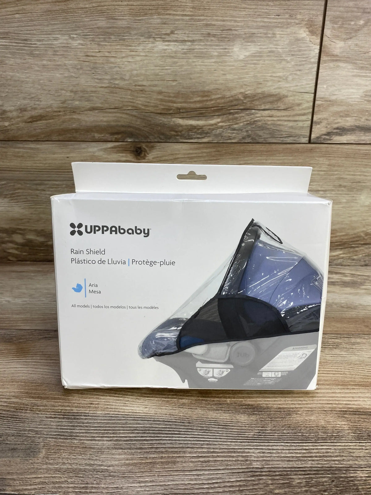 Front view of UPPAbaby Rain Shield for Aria and Mesa car seats showing side vents in packaging on wood floor