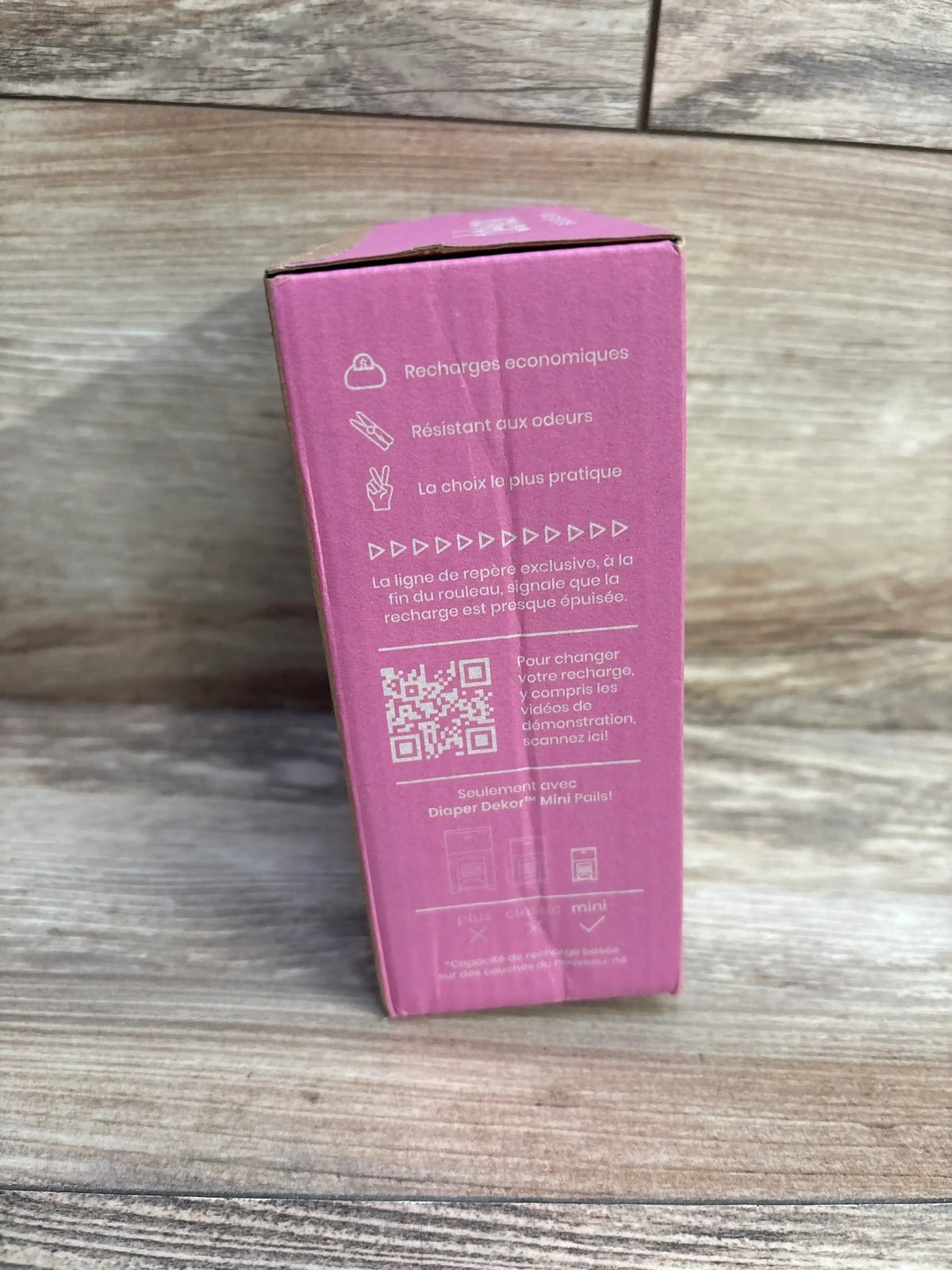Side view of Diaper Dekor Eko Mini refill 2-pack box showing pink packaging with feature icons, French text, and QR code on a wood surface