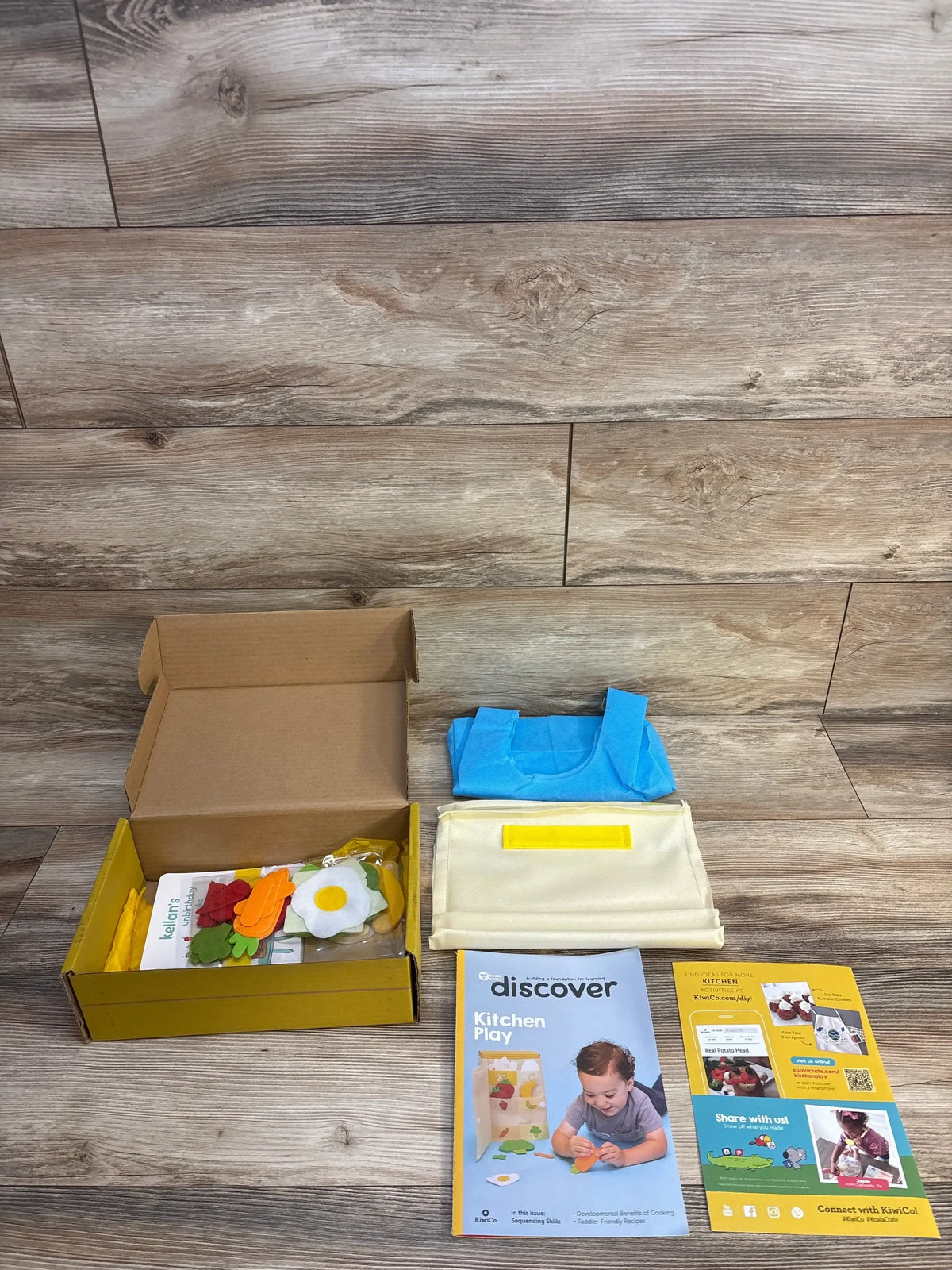 Overhead view of KiwiCo Koala Crate kitchen playset (ages 2+) with felt food, blue apron, and activity booklet on wood floor