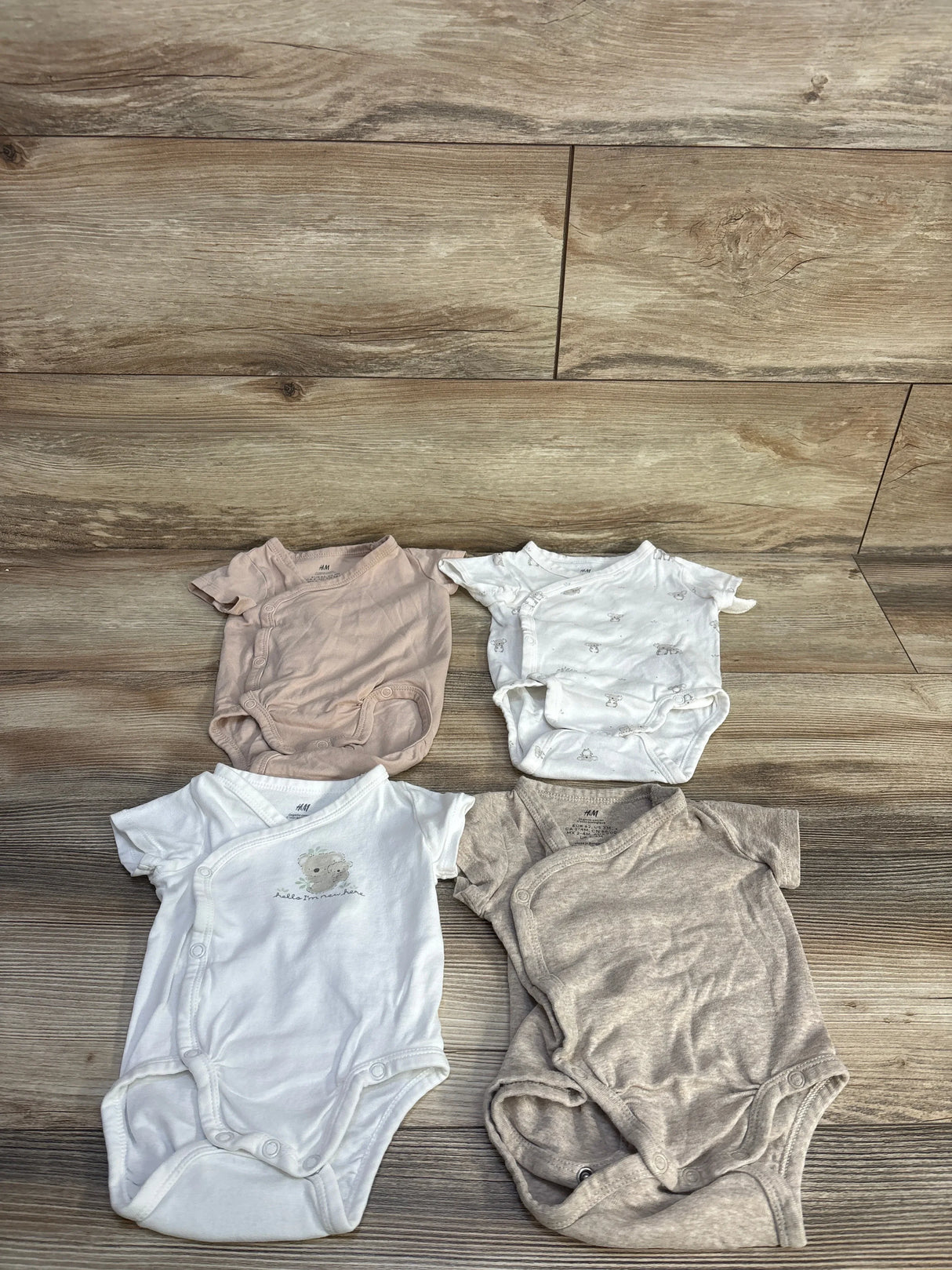 Top-down of four H&M organic kimono baby bodysuits (3M) showing wrap-front snap closures in beige and white on wood floor