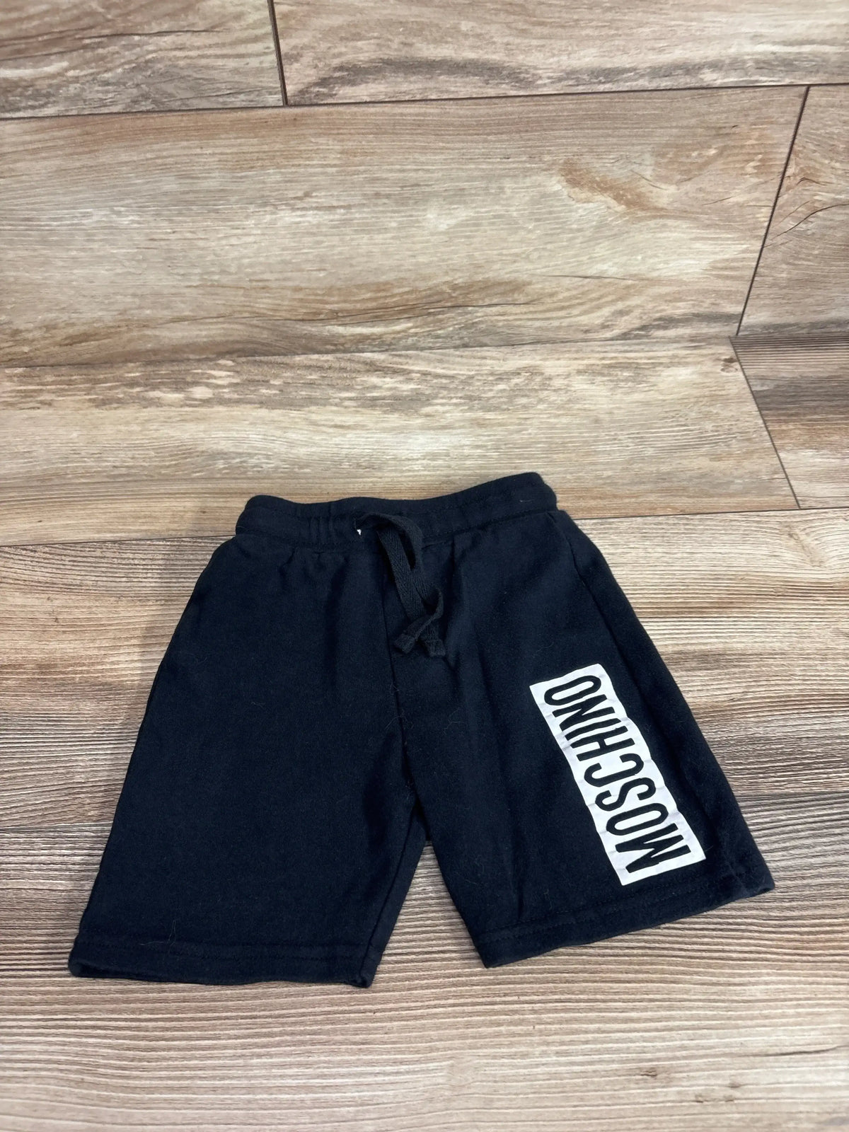 Flat lay of black Moschino baby shorts (12 months) showing vertical white MOSCHINO logo on left leg on wood floor background
