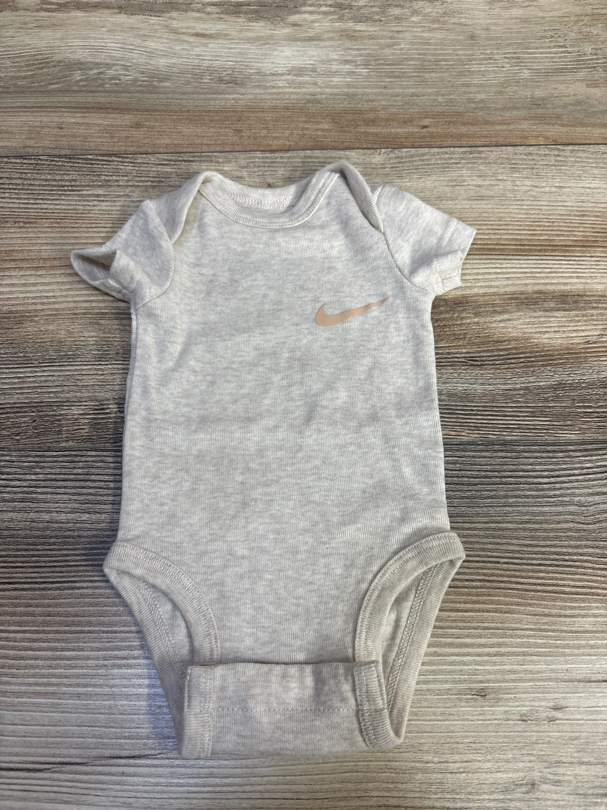 Front view of beige Nike newborn bodysuit showcasing short sleeves and Nike swoosh on a wood-grain background