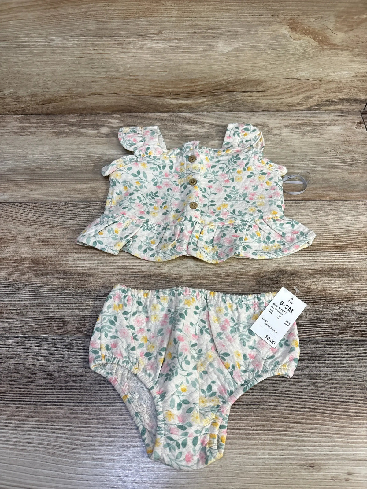 Flat-lay front view of Ivory Cloud Island newborn floral peplum top showcasing ruffled hem and wooden buttons with matching bloomers on wood background