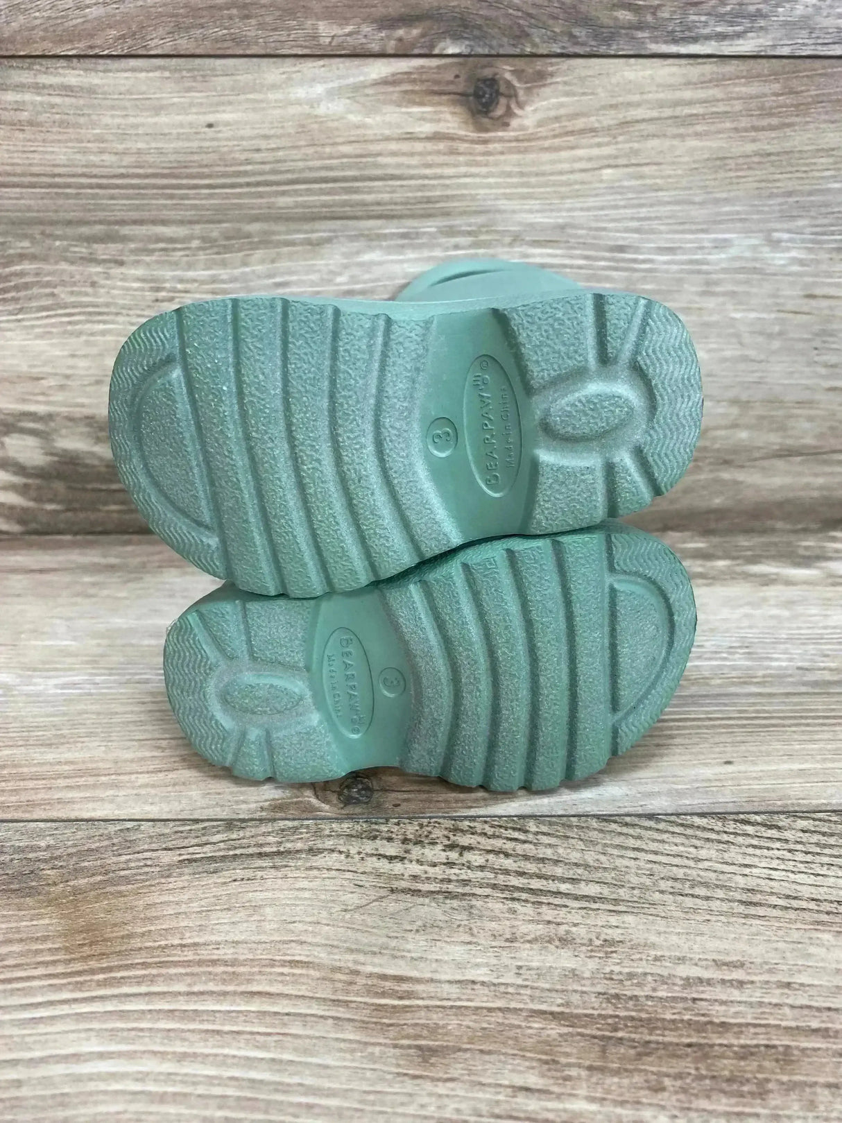 Bottom view of BEARPAW toddler green EVA rain boots showing textured tread, size '3' marking on wood plank background