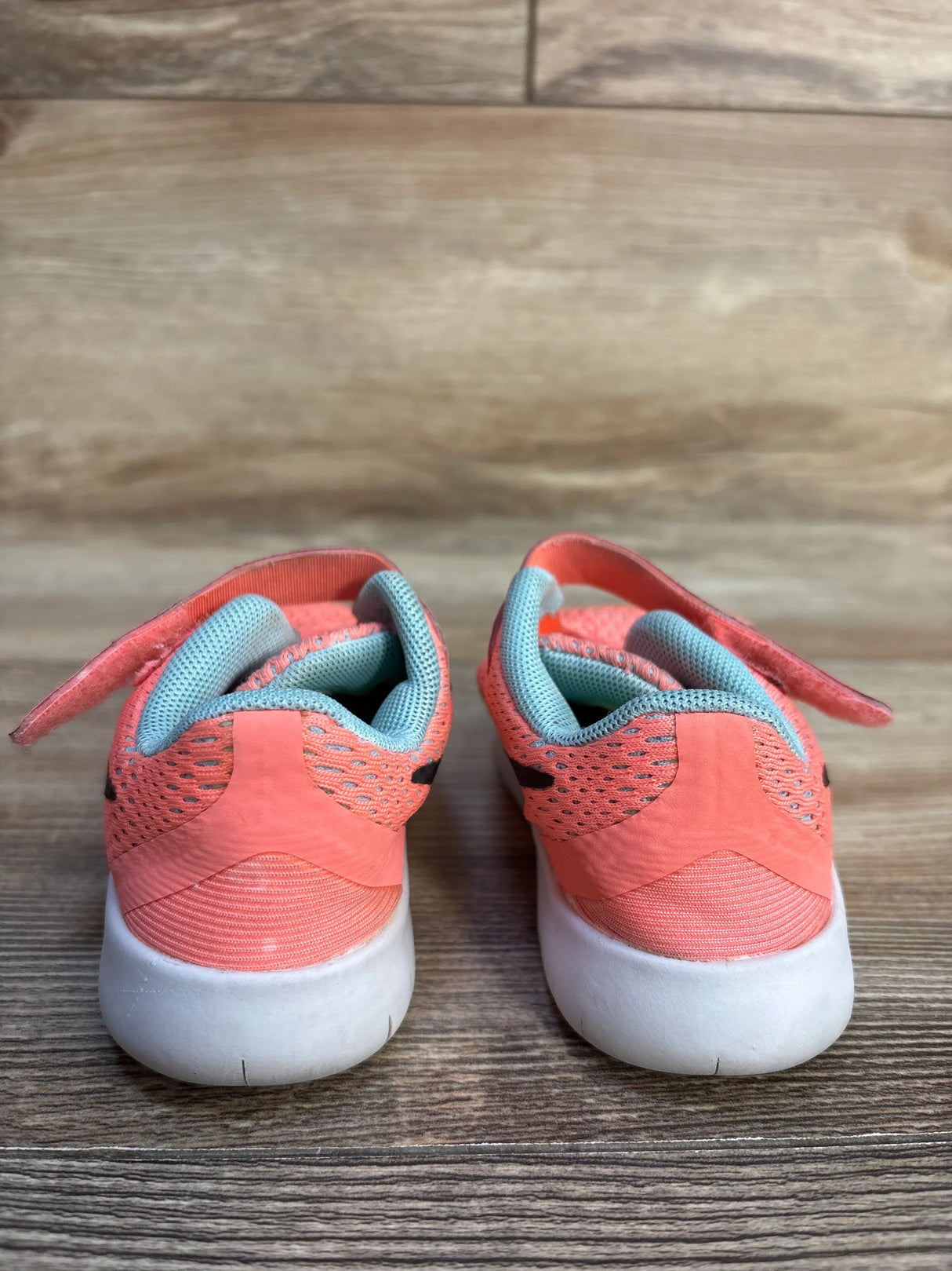 Rear view of kids coral pink mesh sneakers showcasing aqua collar lining and white rubber soles on wood plank floor